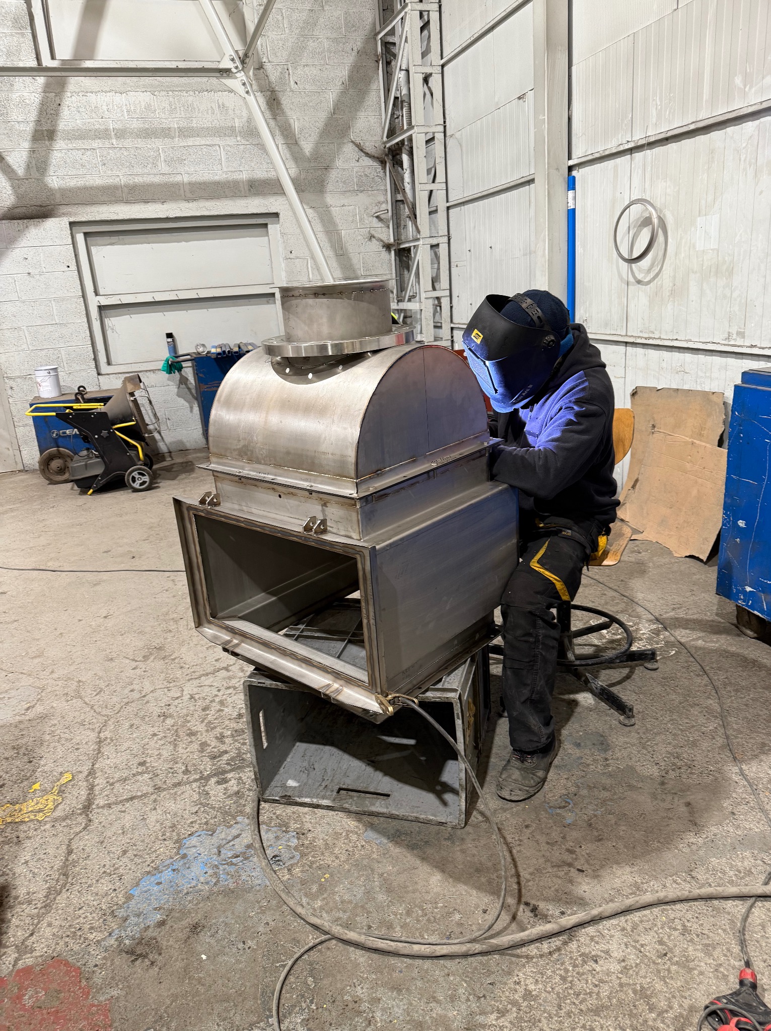 Spawacz w masce spawalniczej łączy metalowe elementy konstrukcji wentylacyjnej w hali, używając spawarki. Widoczne iskry i elementy konstrukcyjne.