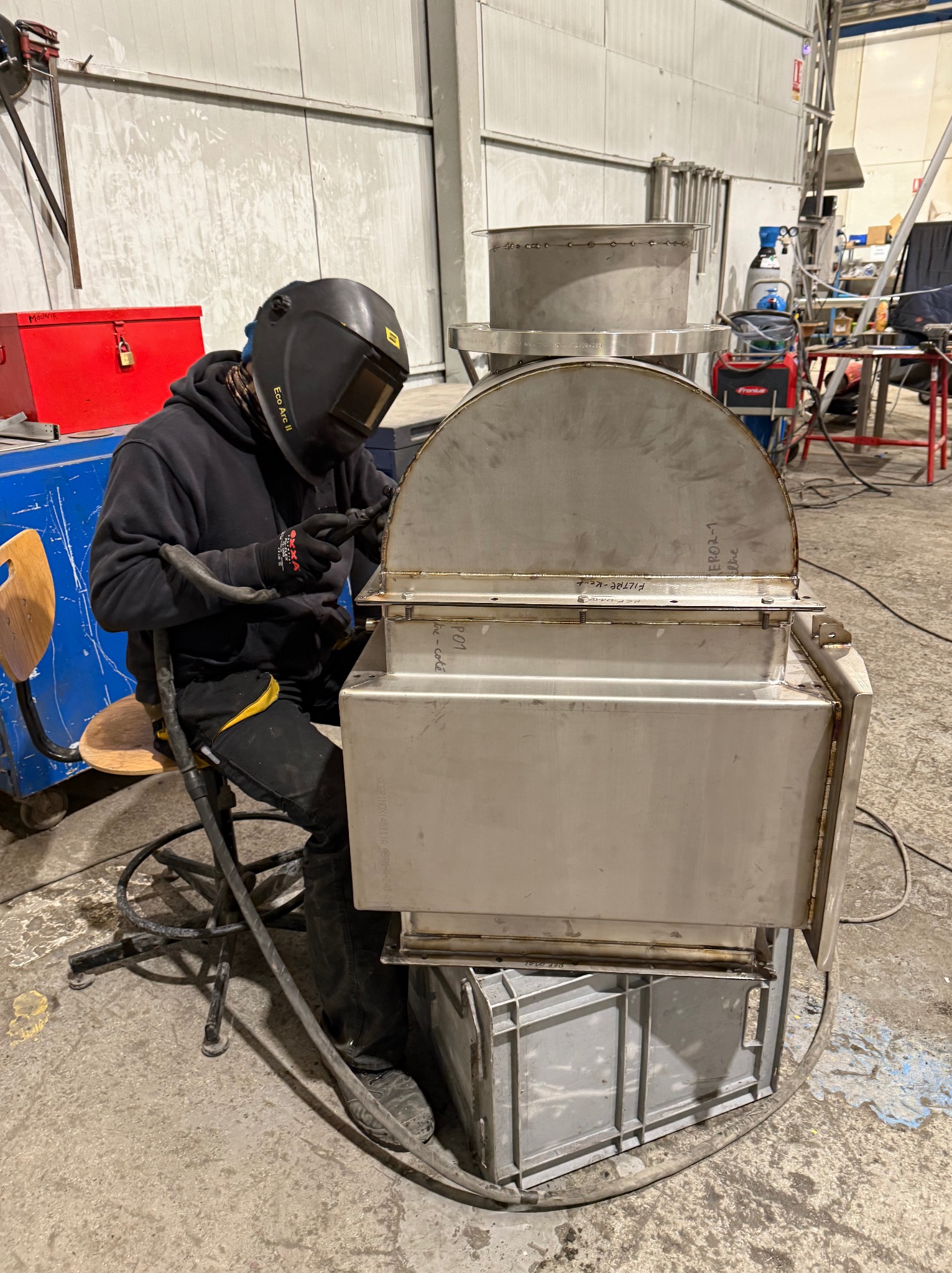 Spawacz w czarnym kombinezonie i masce spawalniczej łączy metalowe elementy konstrukcji w hali przemysłowej. Widoczne iskry i spawarka Fronius.