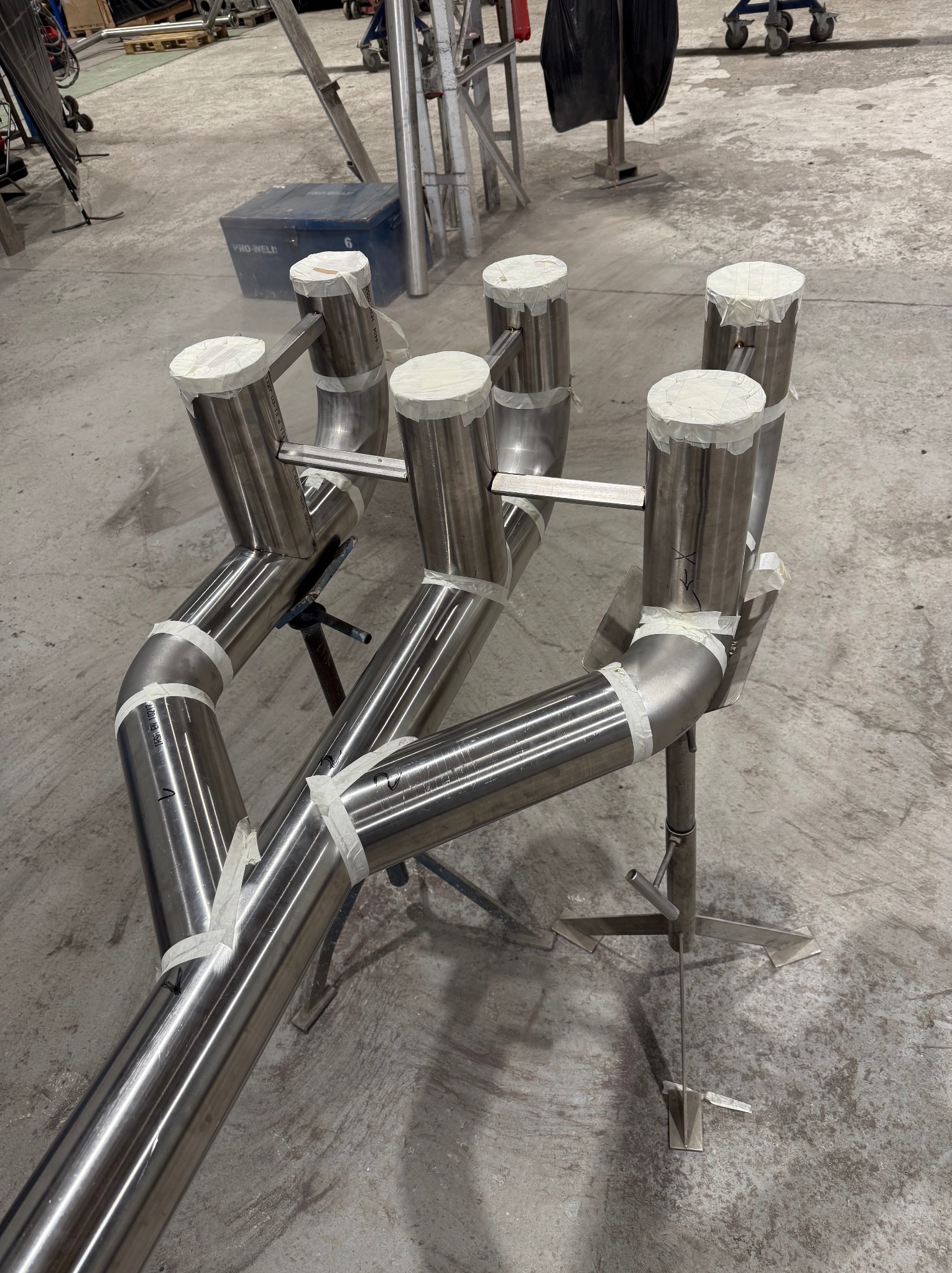 Skomplikowany układ rur ze stali nierdzewnej, połączonych taśmą, na metalowych stojakach, w hali przemysłowej. Widoczne złącza i oznaczenia na rurach.