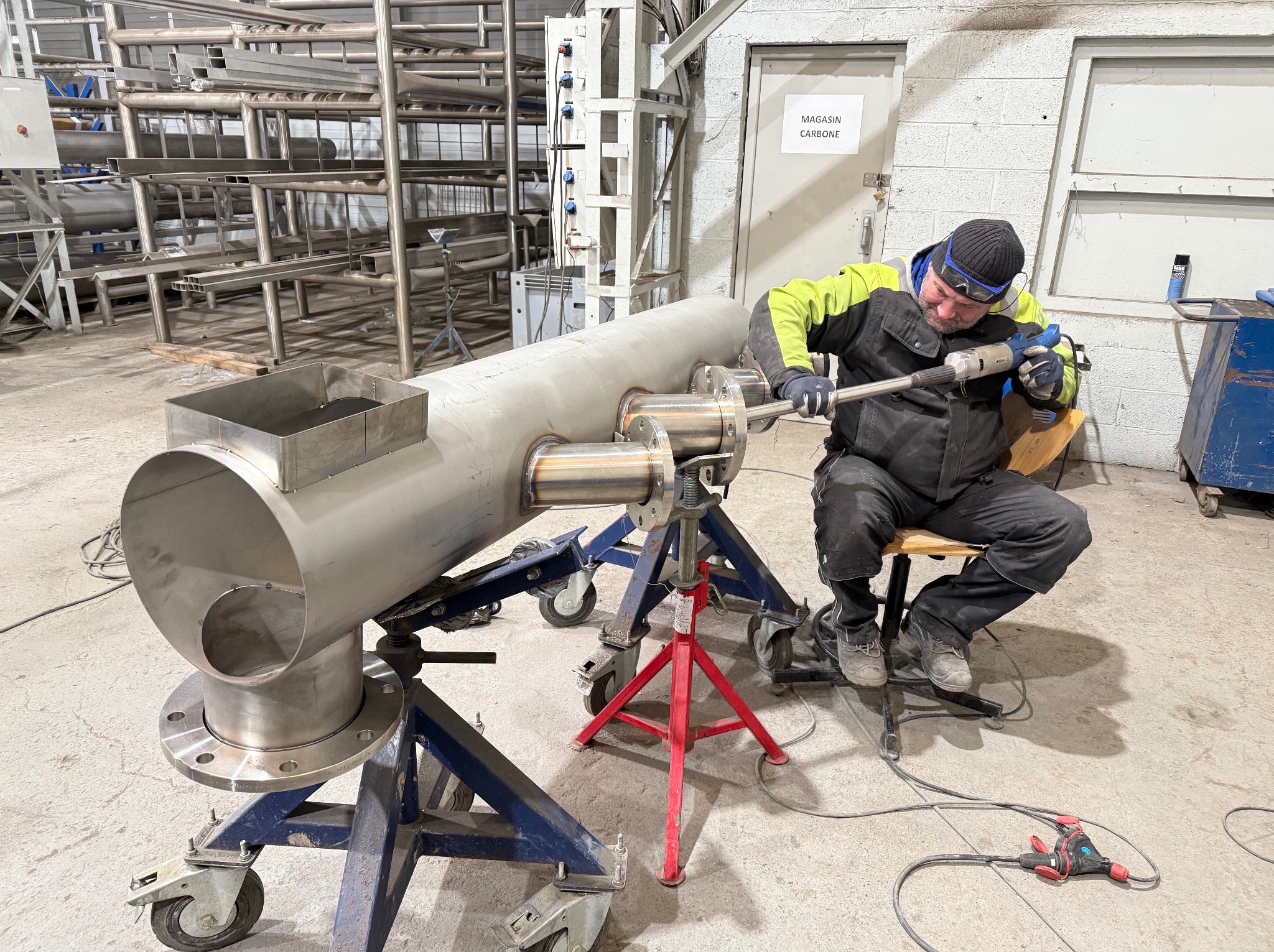 Spawanie rury ze stali nierdzewnej przez montera w warsztacie. Rura na wózku warsztatowym. Widoczne narzędzia i wyposażenie. Magazyn Carbone w tle.