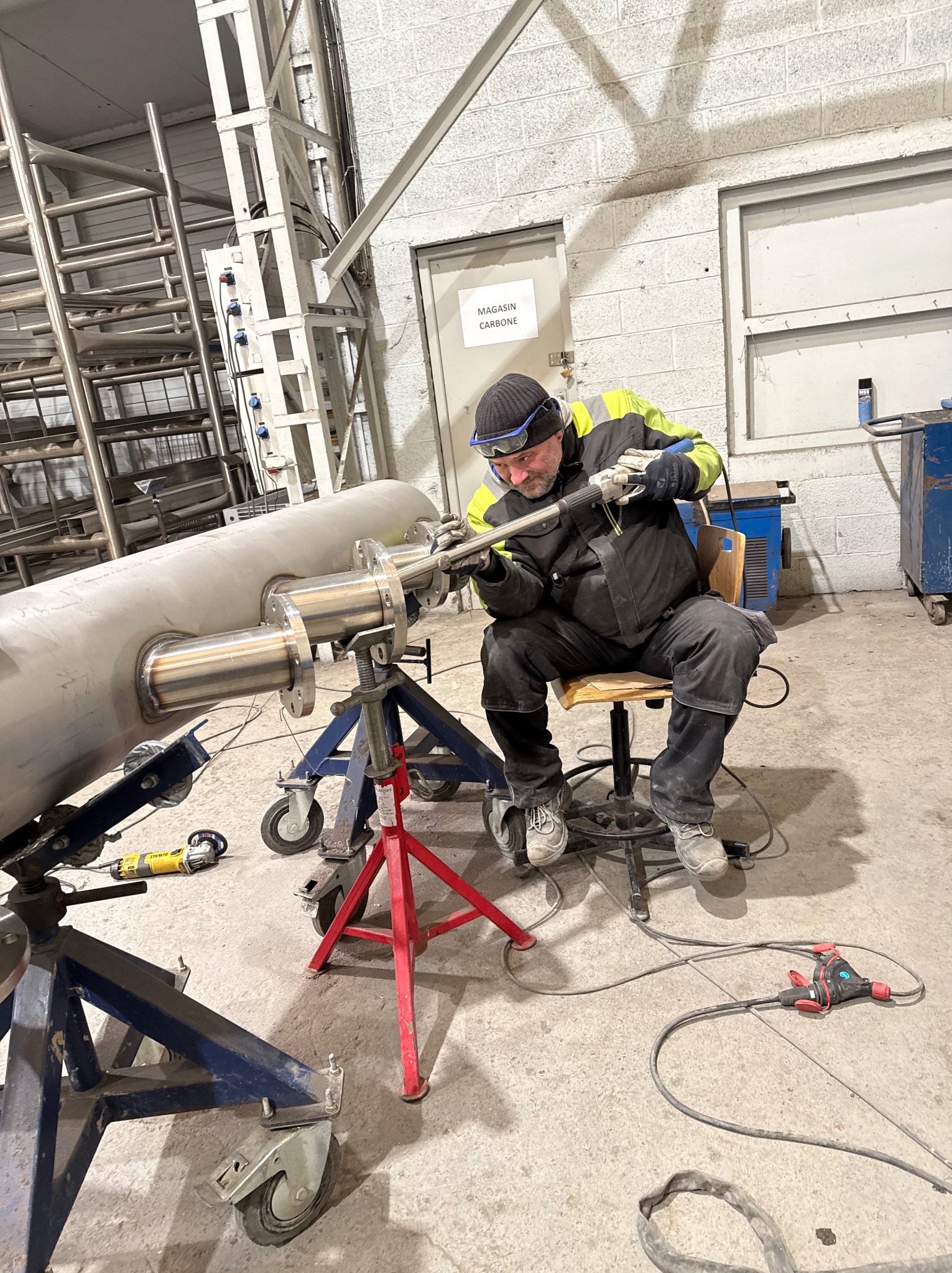 Spawacz w Szczytnie łączy rury ze stali nierdzewnej w hali produkcyjnej, używając profesjonalnego sprzętu spawalniczego. Widoczne zabezpieczenia i narzędzia.