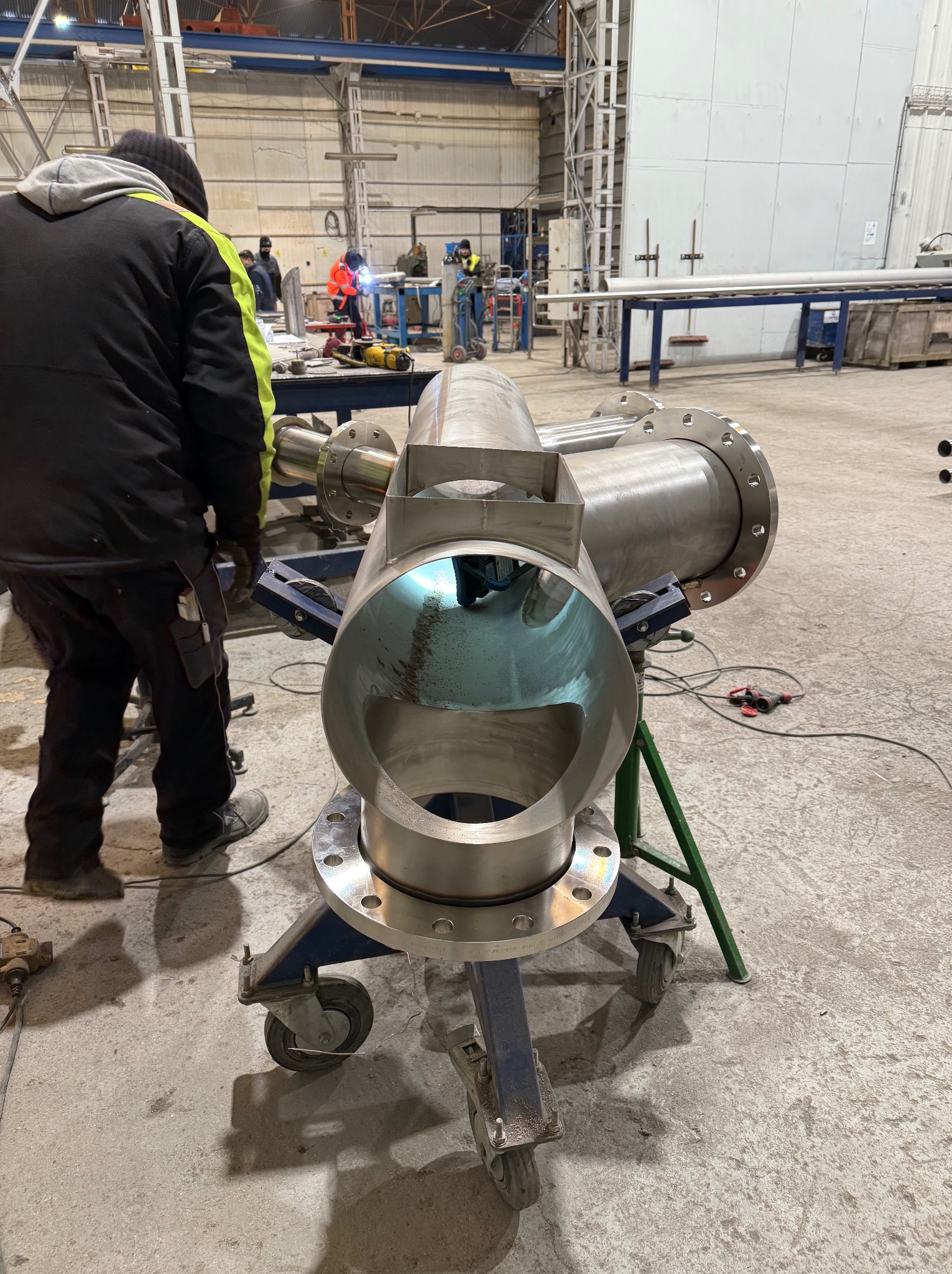 Spawanie rur ze stali nierdzewnej na wózku transportowym w hali produkcyjnej. Widoczni spawacze w tle, narzędzia i elementy konstrukcyjne.