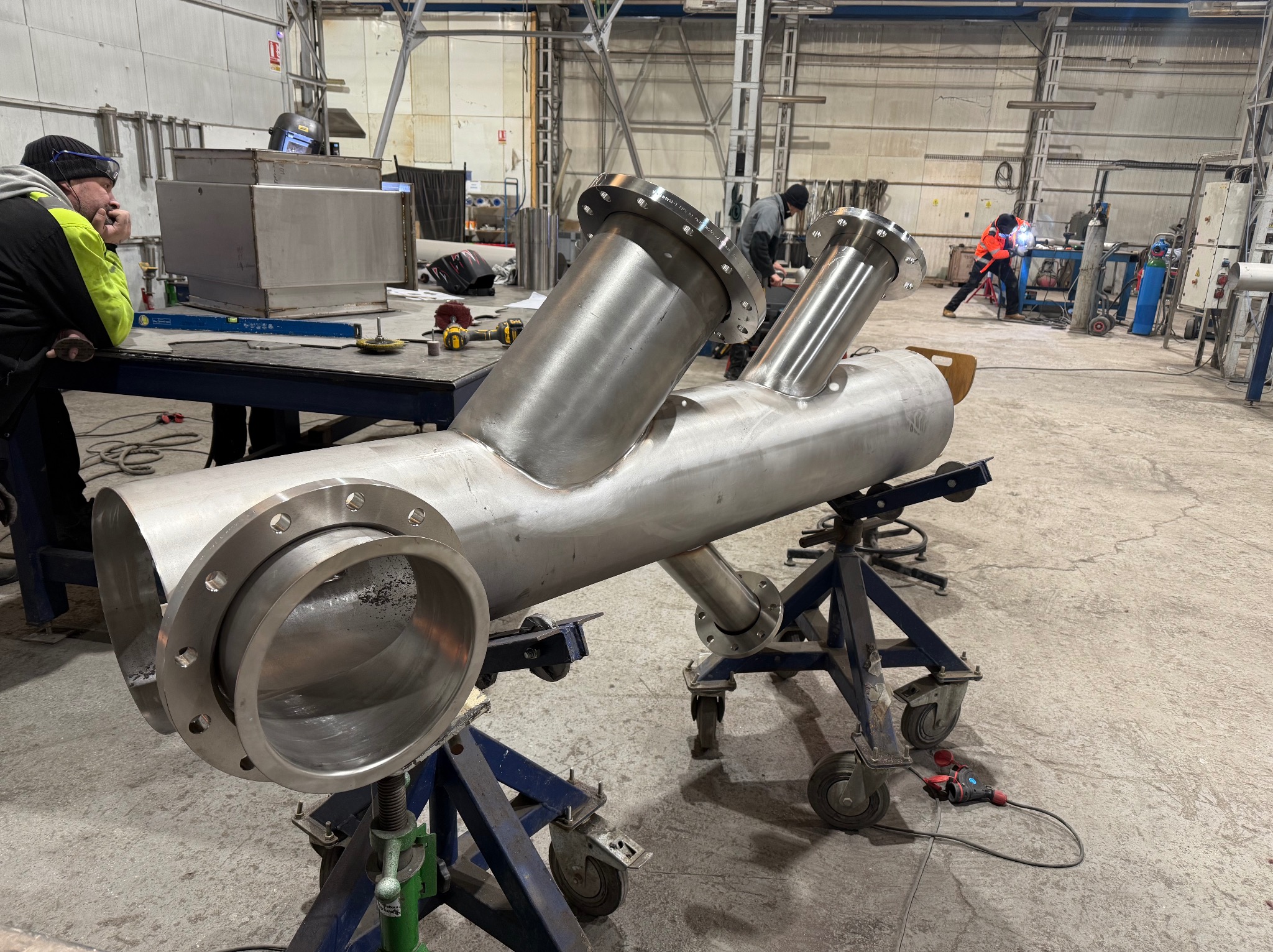 Spawana, nierdzewna konstrukcja rurowa z kołnierzami na wózkach warsztatowych. W tle spawacz w kombinezonie i inne elementy wyposażenia hali.