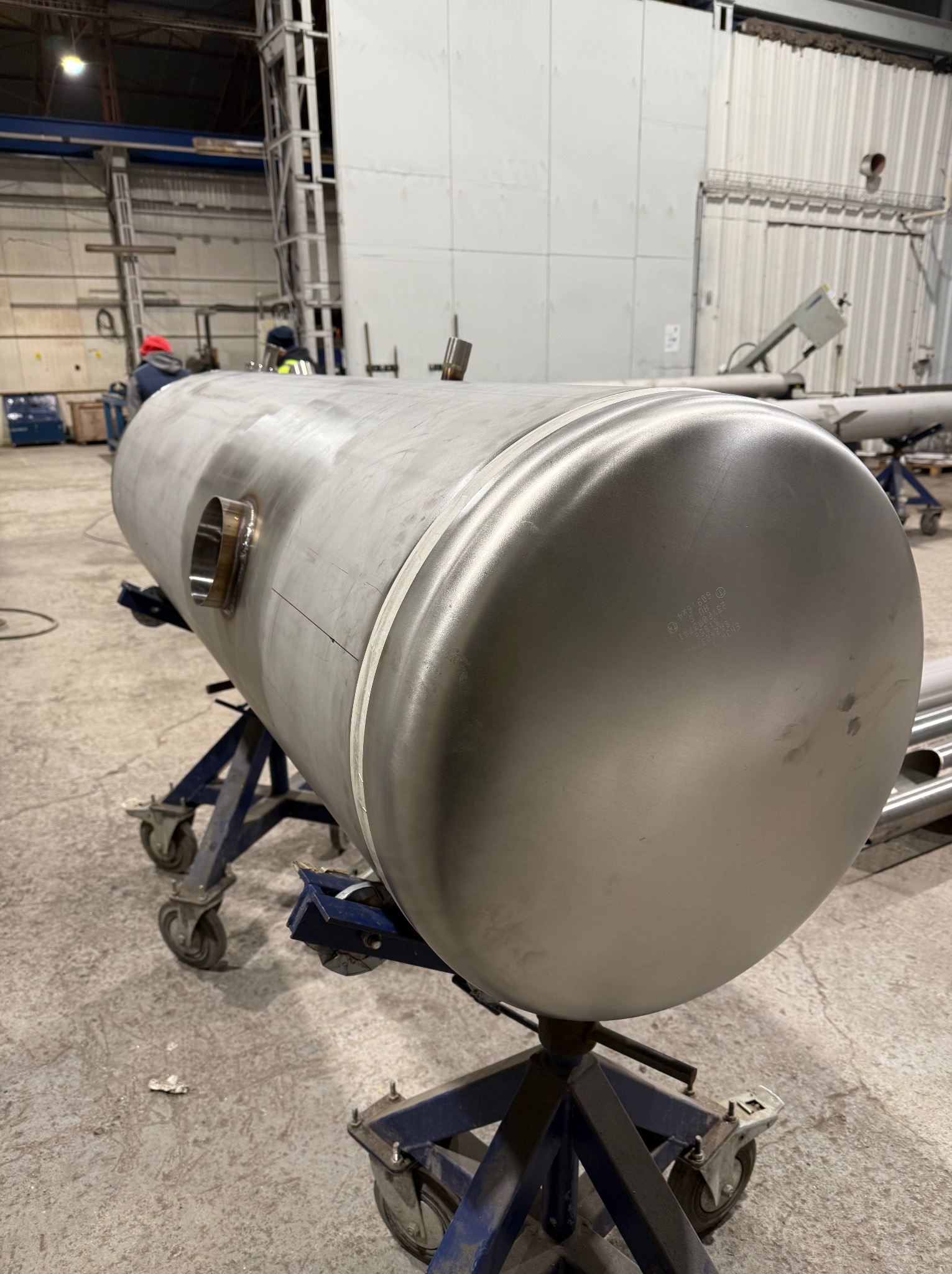 Metalowy, cylindryczny zbiornik na niebieskim wózku, z widocznym spawem i otworem. W tle hala produkcyjna z pracownikami i konstrukcjami stalowymi.