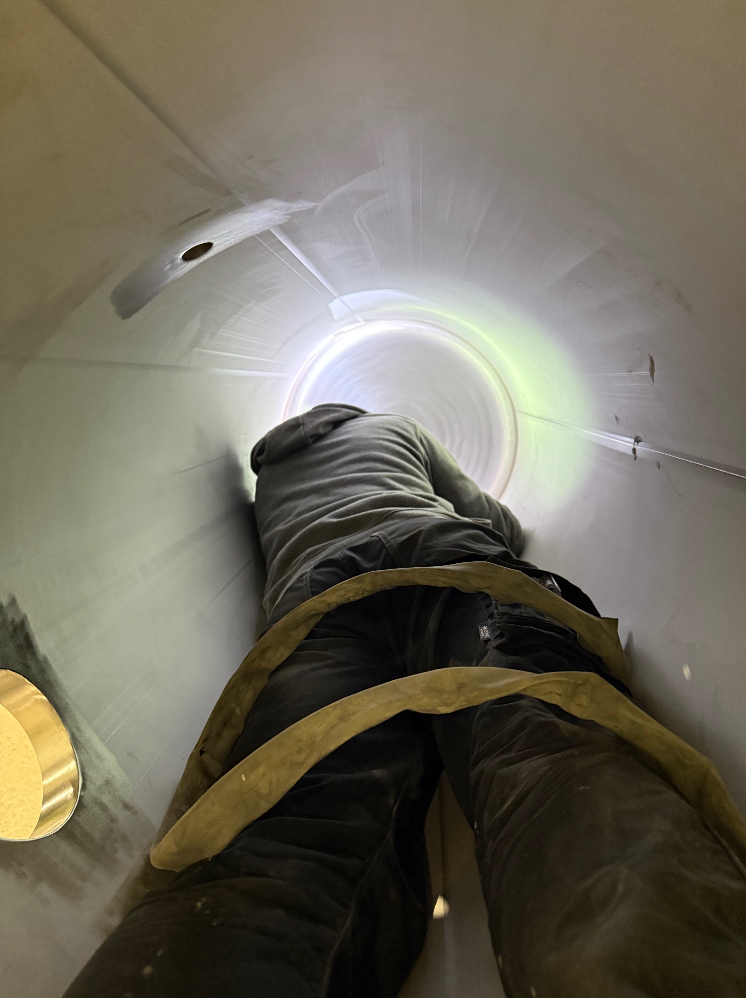 Pracownik wewnątrz metalowej rury o dużej średnicy, prawdopodobnie komina lub kanału wentylacyjnego, w trakcie inspekcji lub prac konserwacyjnych. Widok z dołu.