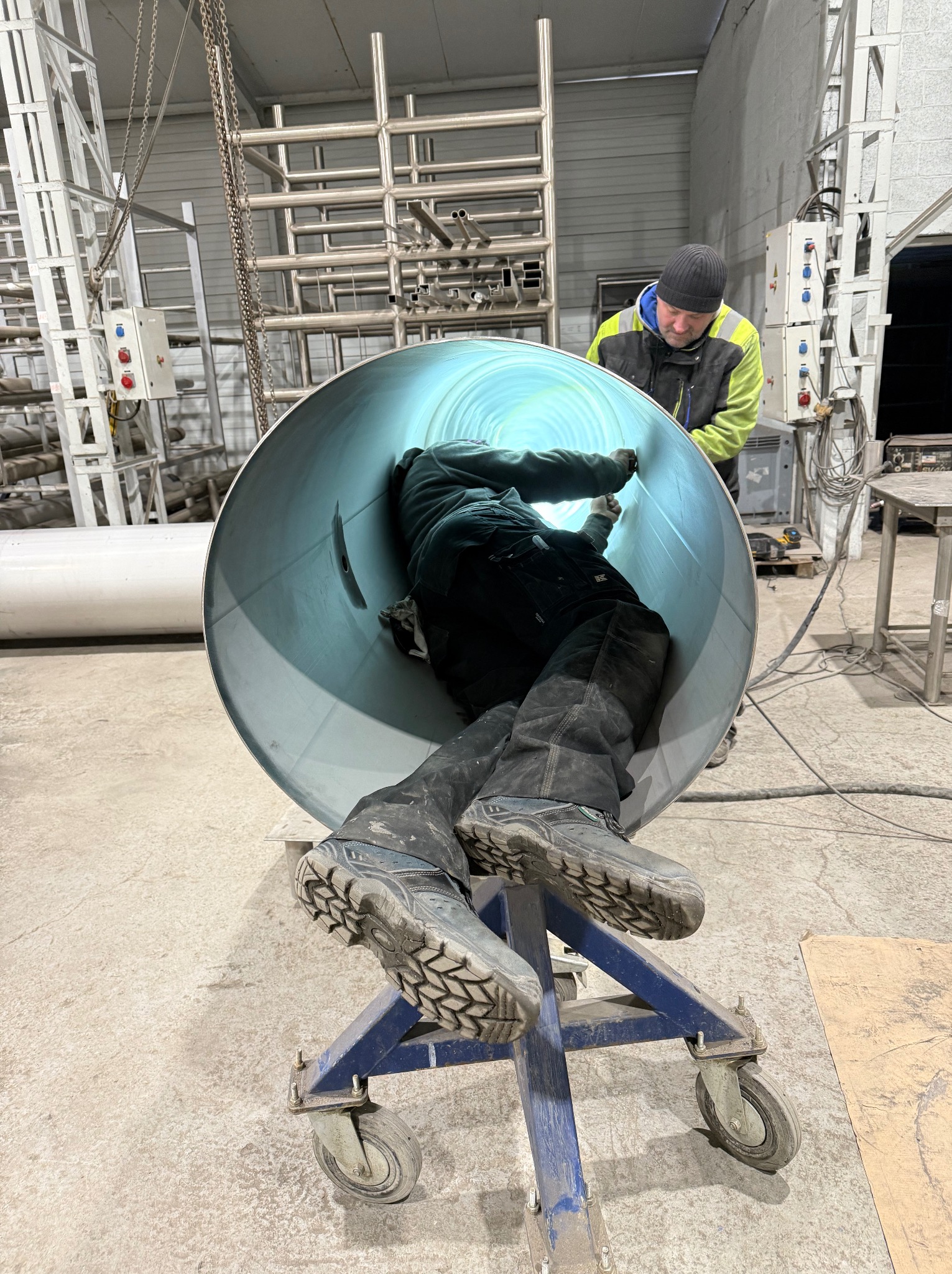 Pracownik wewnątrz dużej, jasnej rury na wózku, drugi pracownik nadzoruje. Hala przemysłowa w tle, widoczne instalacje i urządzenia. Nietypowy kadr.