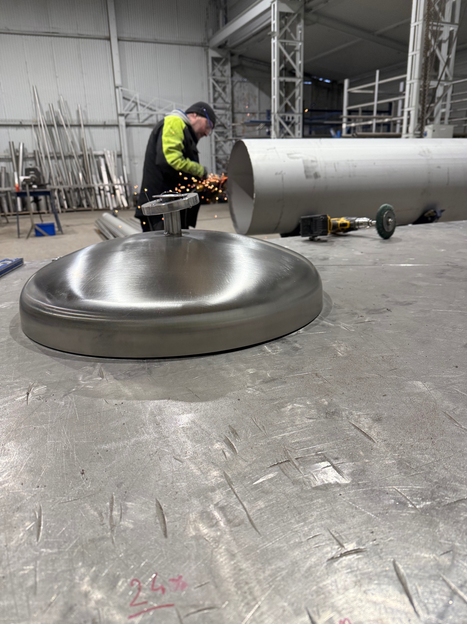 Spawacz w hali obrabia metalową rurę szlifierką kątową. Na pierwszym planie polerowana metalowa pokrywa. W tle rury i konstrukcja hali.