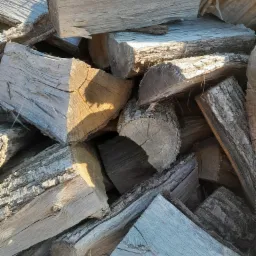 Stos ułożonego drewna kominkowego w różnych odcieniach szarości i brązu, z widoczną korą i śladami cięcia, oświetlony z góry, tworzący kontrast cieni.