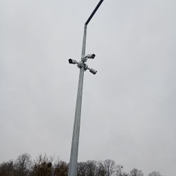 PHU OLSZNET Systemy Zabezpieczeń - Srebrny słup oświetleniowy z zamontowanymi czterema kamerami monitoringu, skierowanymi w różne strony, na tle pochmurnego nieba i drzew bez liści.