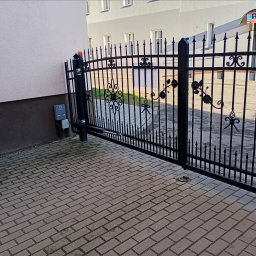 PHU OLSZNET Systemy Zabezpieczeń - Otwarta, czarna, metalowa brama z automatyką na posesji. Widoczny napęd i sterowanie. Kostka brukowa na podjeździe. Budynek w tle.