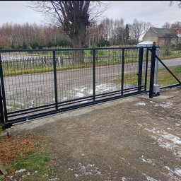 PHU OLSZNET Systemy Zabezpieczeń - Czarna, przesuwna brama z siatki na posesji, z widocznym mechanizmem automatyki. W tle drzewa i budynek. Ujęcie z bliska, podkreślające detale montażu.