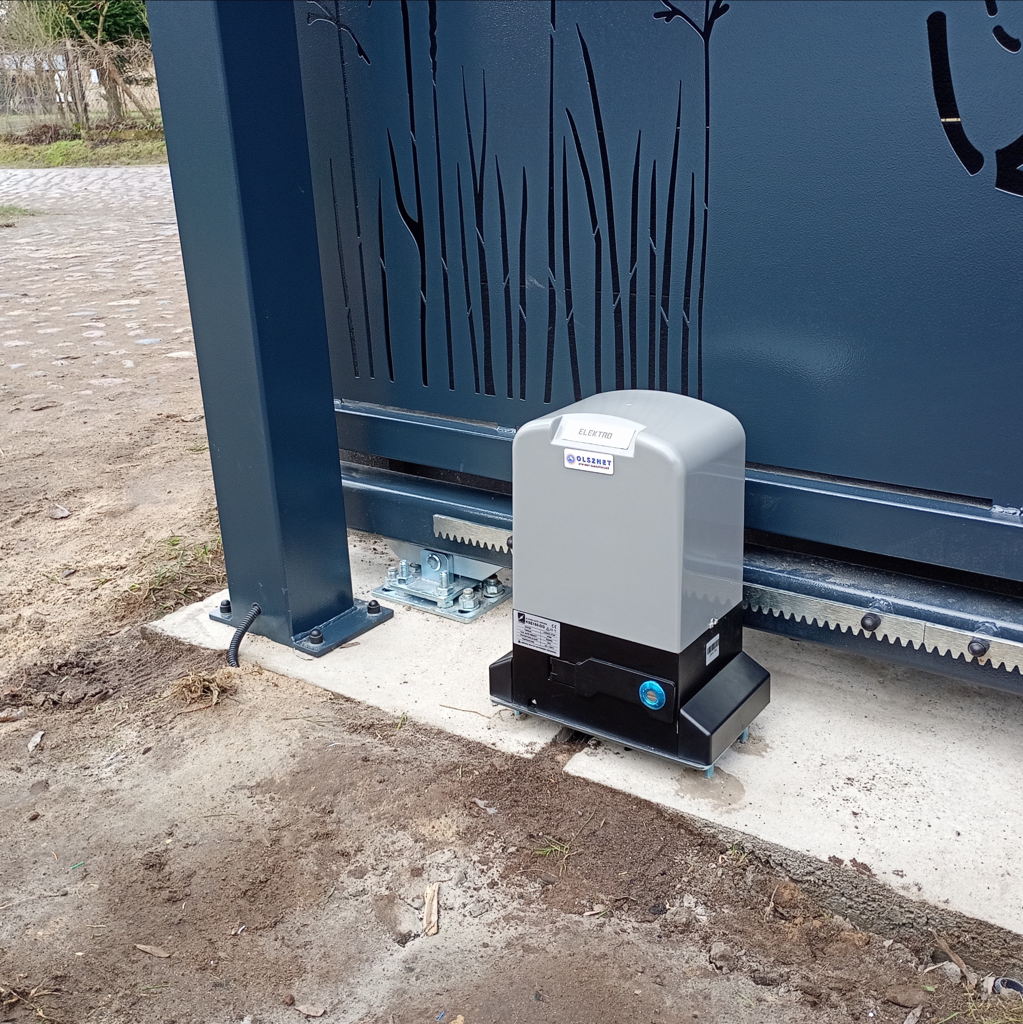 Szary napęd elektryczny do bramy przesuwnej z zębatą listwą, zamontowany na betonowej podstawie obok ozdobnego, ażurowego ogrodzenia w kolorze grafitowym.