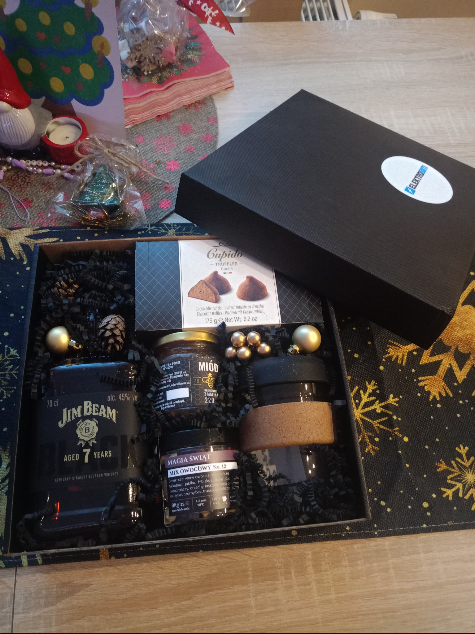 Otwarty, czarny prezentowy box z alkoholem Jim Beam, miodem, truflami i ozdobami świątecznymi na drewnianym stole, obok kartka z choinką i logo firmy.