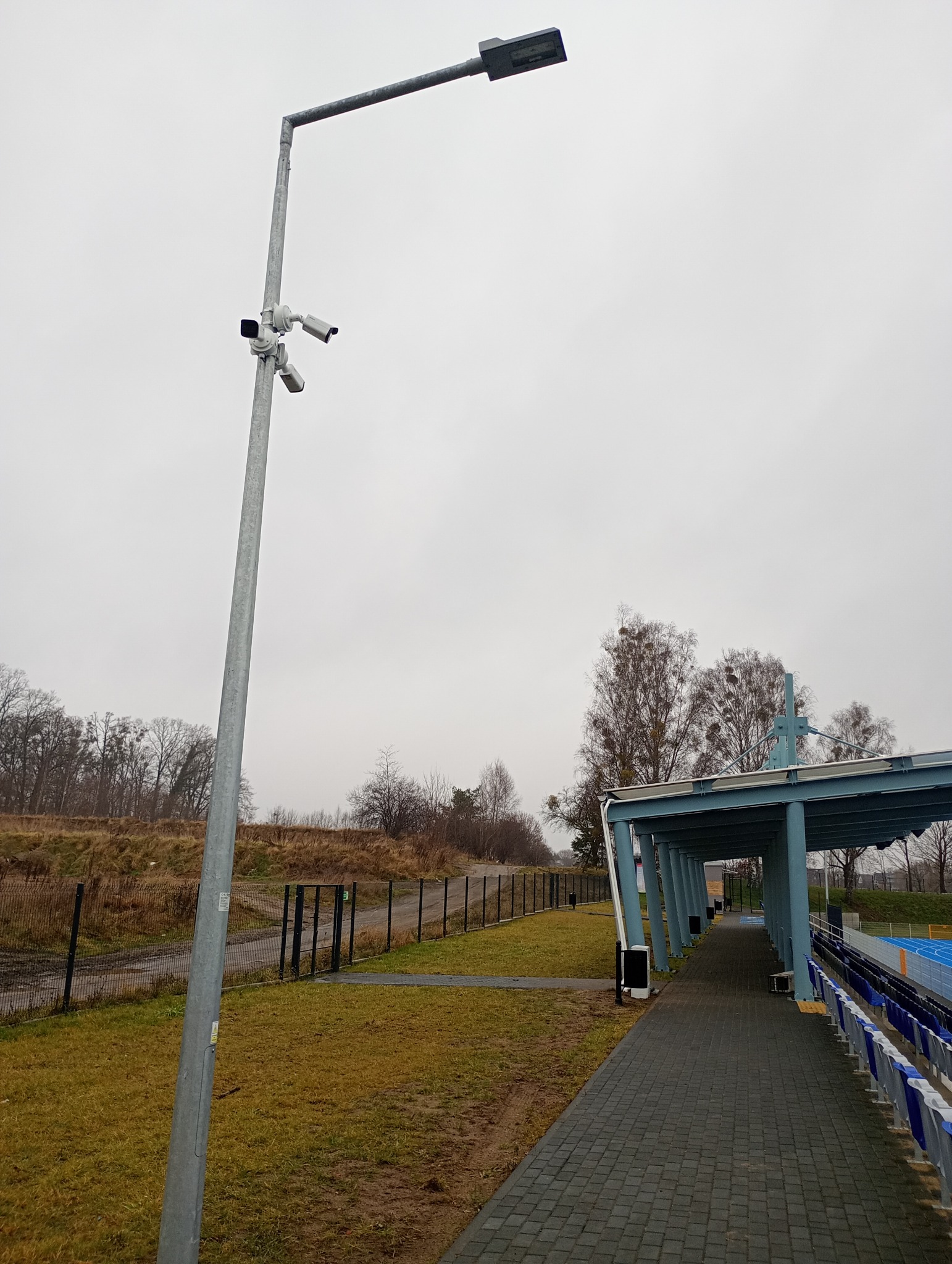 Słup oświetleniowy z trzema kamerami monitoringu na tle pochmurnego nieba, obok trybun stadionu. Widok na zainstalowany system monitoringu.