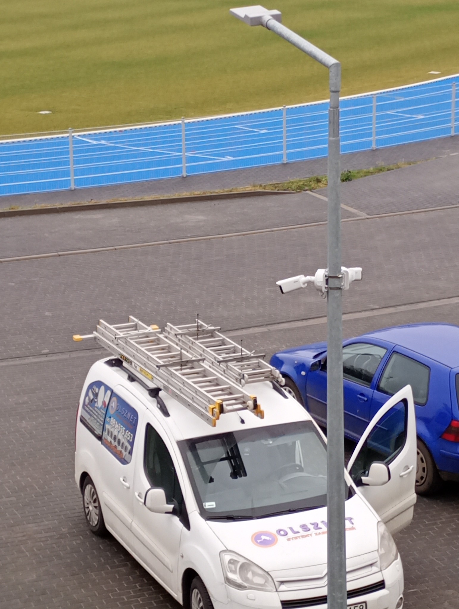 Biały samochód serwisowy z drabiną na dachu, zaparkowany obok słupa z kamerą monitoringu na tle stadionu lekkoatletycznego. Otwarta boczna szyba auta.