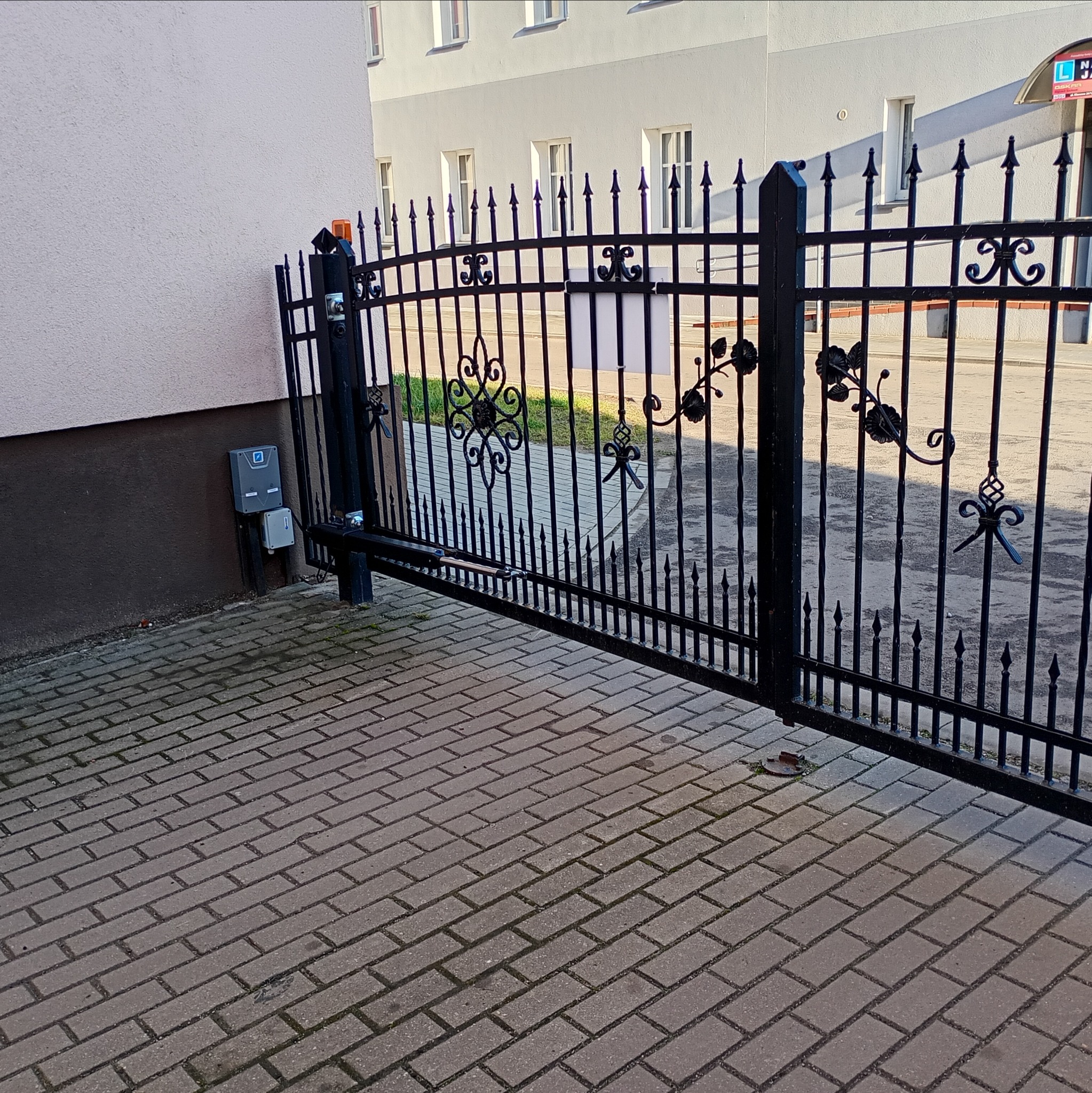 Otwarta, czarna, metalowa brama z automatyką na posesji. Widoczny napęd i sterowanie. Kostka brukowa na podjeździe. Budynek w tle.
