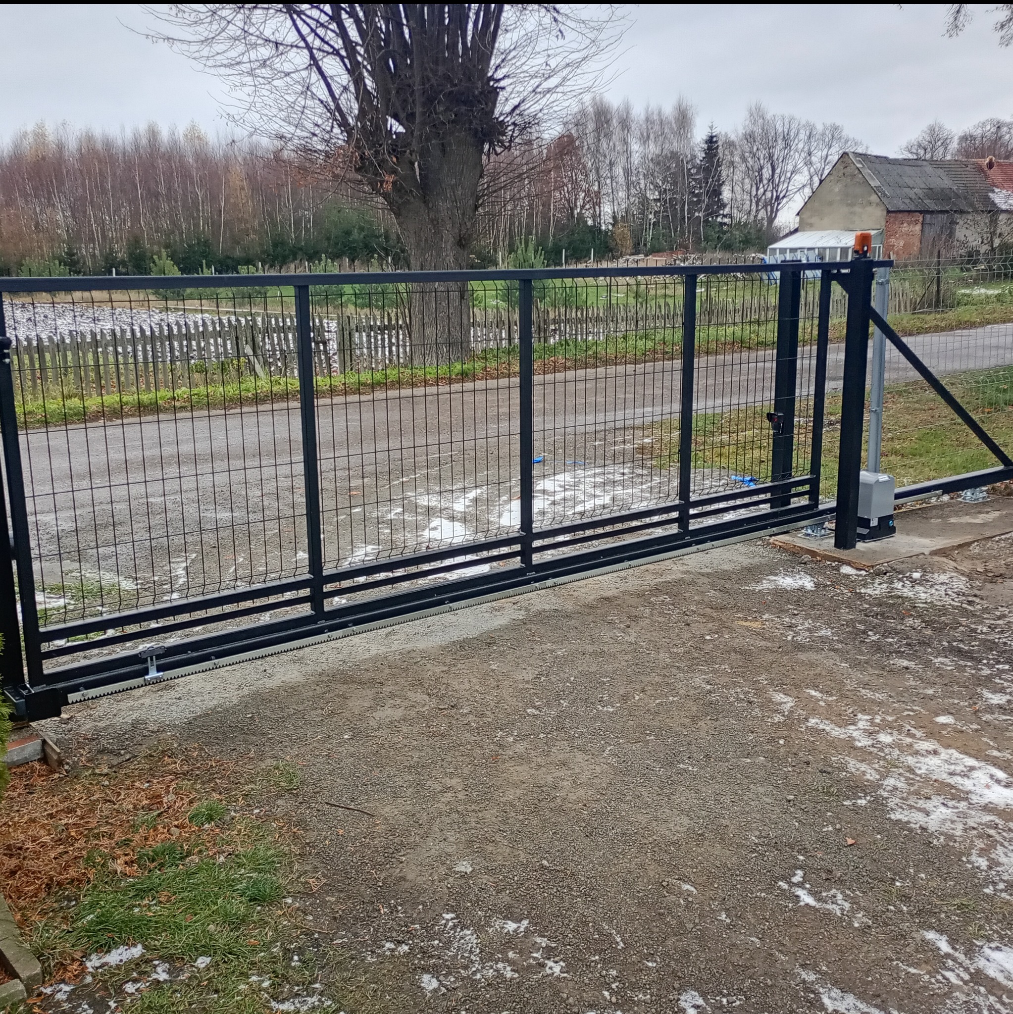 Czarna, przesuwna brama z siatki na posesji, z widocznym mechanizmem automatyki. W tle drzewa i budynek. Ujęcie z bliska, podkreślające detale montażu.