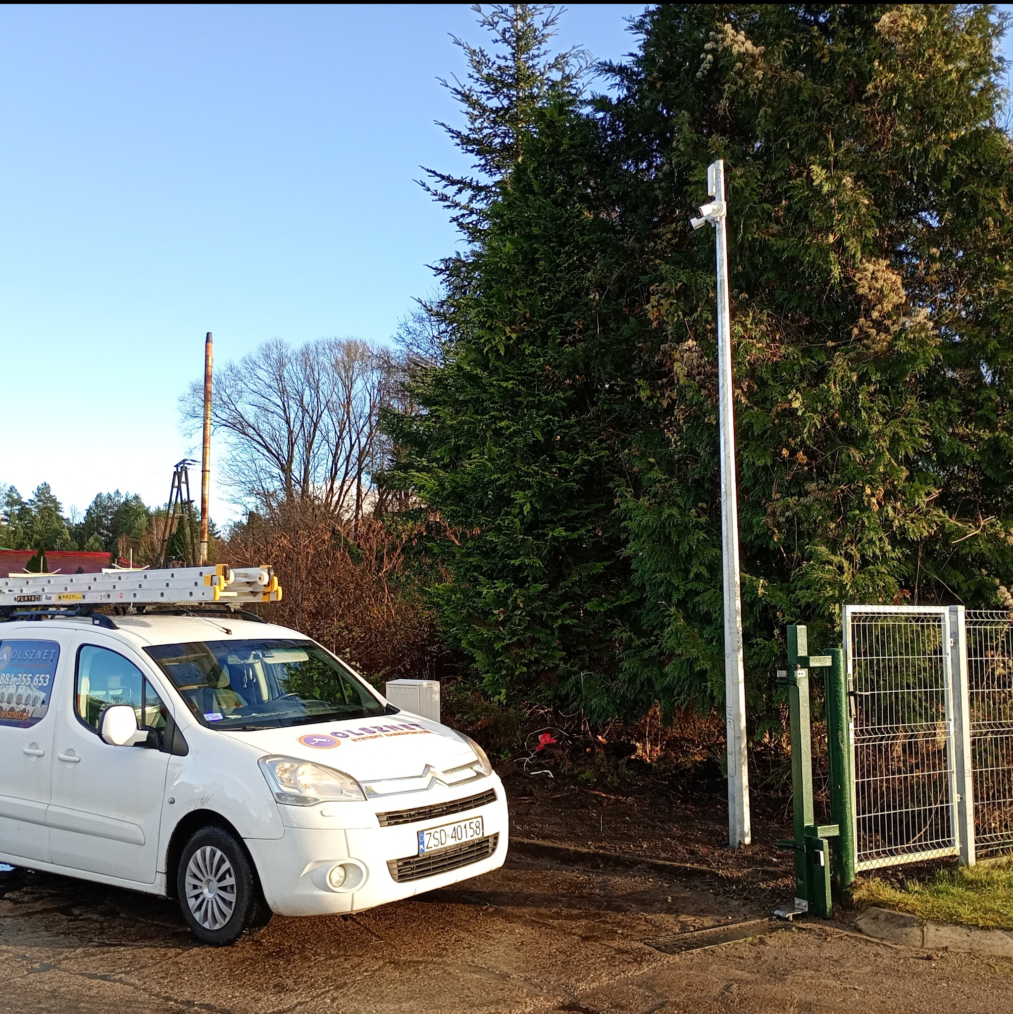 Biały samochód z drabiną na dachu zaparkowany obok słupa z kamerami monitoringu przy ogrodzeniu. W tle drzewa i komin.