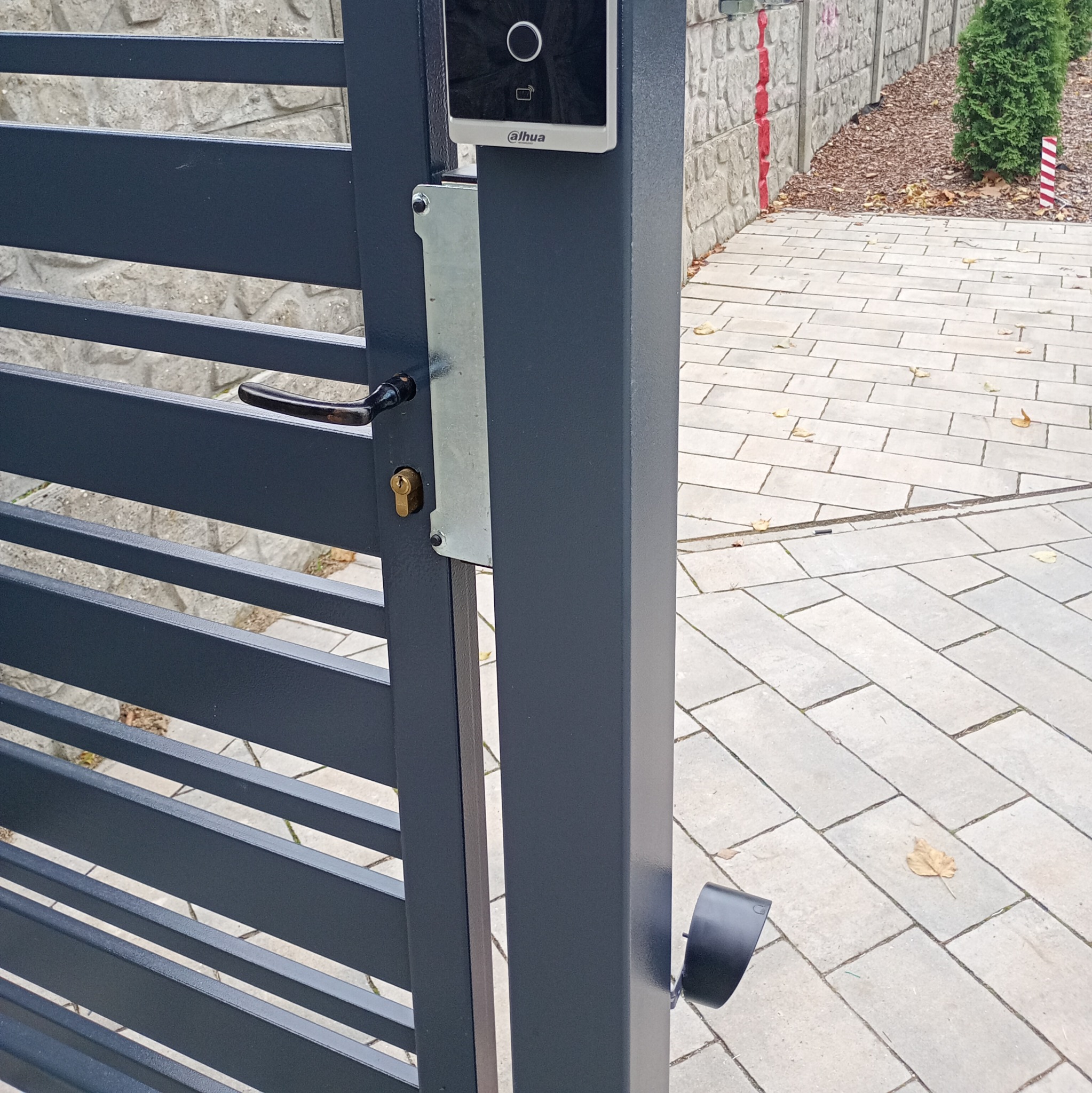 Nowoczesna, grafitowa brama z zamontowanym wideodomofonem Dahua. Widoczny fragment chodnika z kostki i ogrodzenie z kamienia.