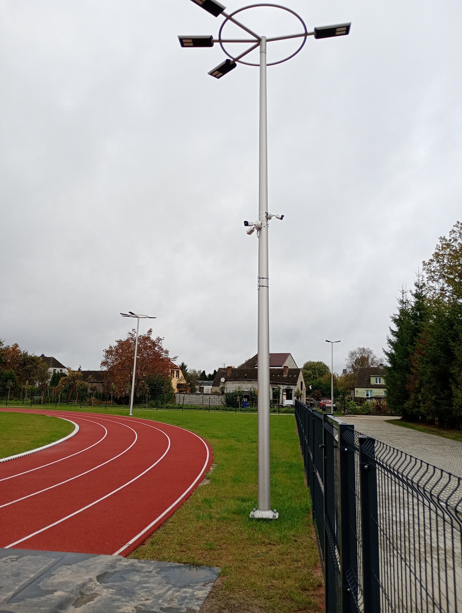 Oświetlenie i monitoring boiska sportowego: wysoki słup z reflektorami LED i kamerami przemysłowymi, widok na bieżnię i ogrodzenie panelowe. Pochmurne niebo w tle.