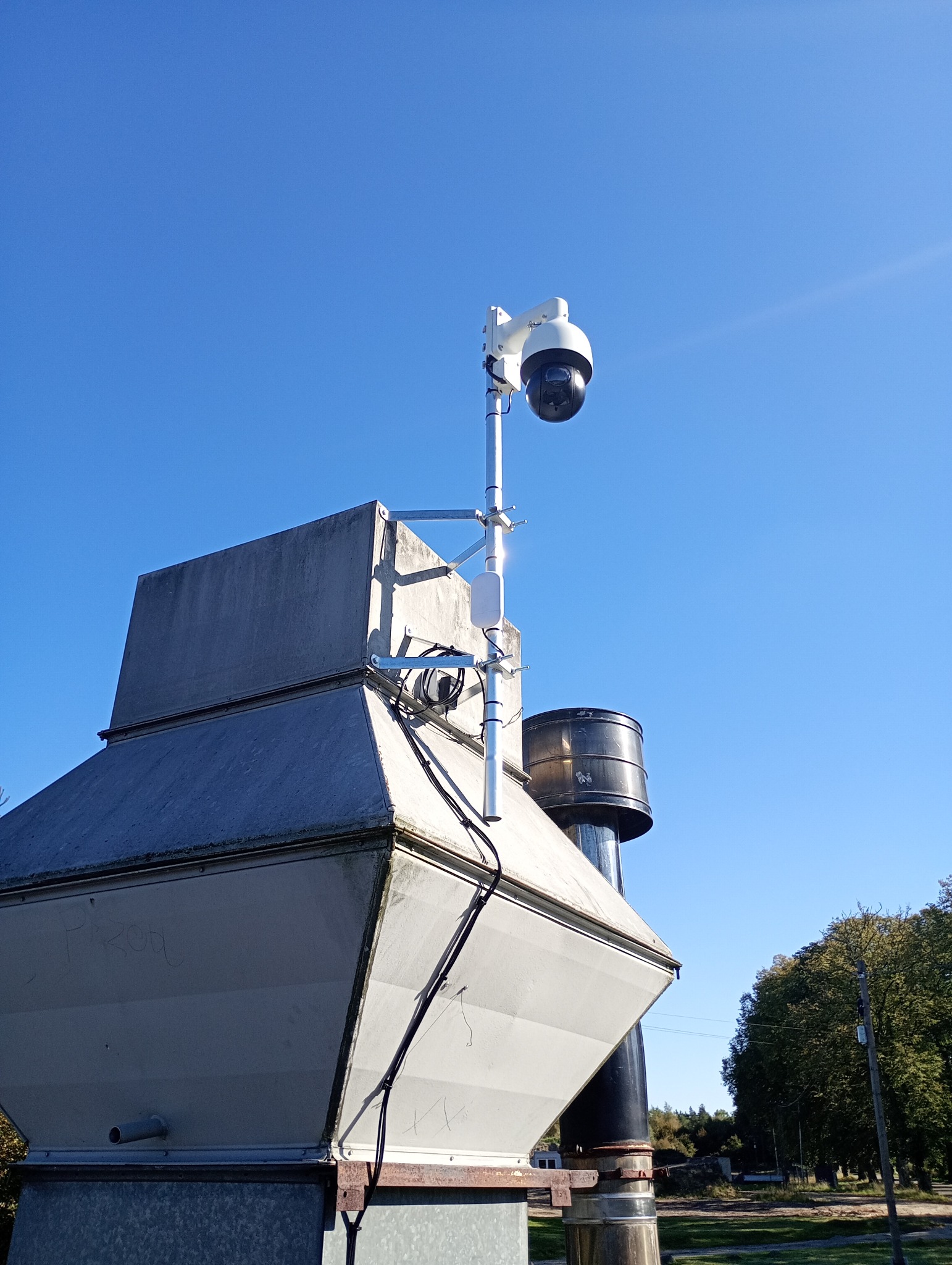 Kamera monitoringu PTZ zamontowana na dachu budynku przemysłowego, widoczna instalacja kablowa i elementy mocujące. Błękitne niebo w tle.