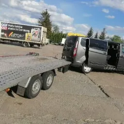 Szary van z otwartymi drzwiami wciągany na przyczepę lawetę, w tle ciężarówka z reklamą olejów silnikowych.