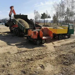 Ciężarówka z dźwigiem załadowuje rozdrobnione gałęzie do kontenera, obok stoi rozdrabniacz do gałęzi i mała ciężarówka z żółtą skrzynią ładunkową, teren wokół zniszczony po pracy maszyn.