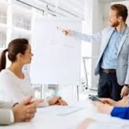 Mężczyzna w jasnej marynarce prezentuje wykres na białej tablicy podczas spotkania biznesowego z zespołem w sali konferencyjnej.