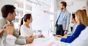 Mężczyzna w jasnej marynarce prezentuje wykres na białej tablicy podczas spotkania biznesowego z zespołem w sali konferencyjnej.