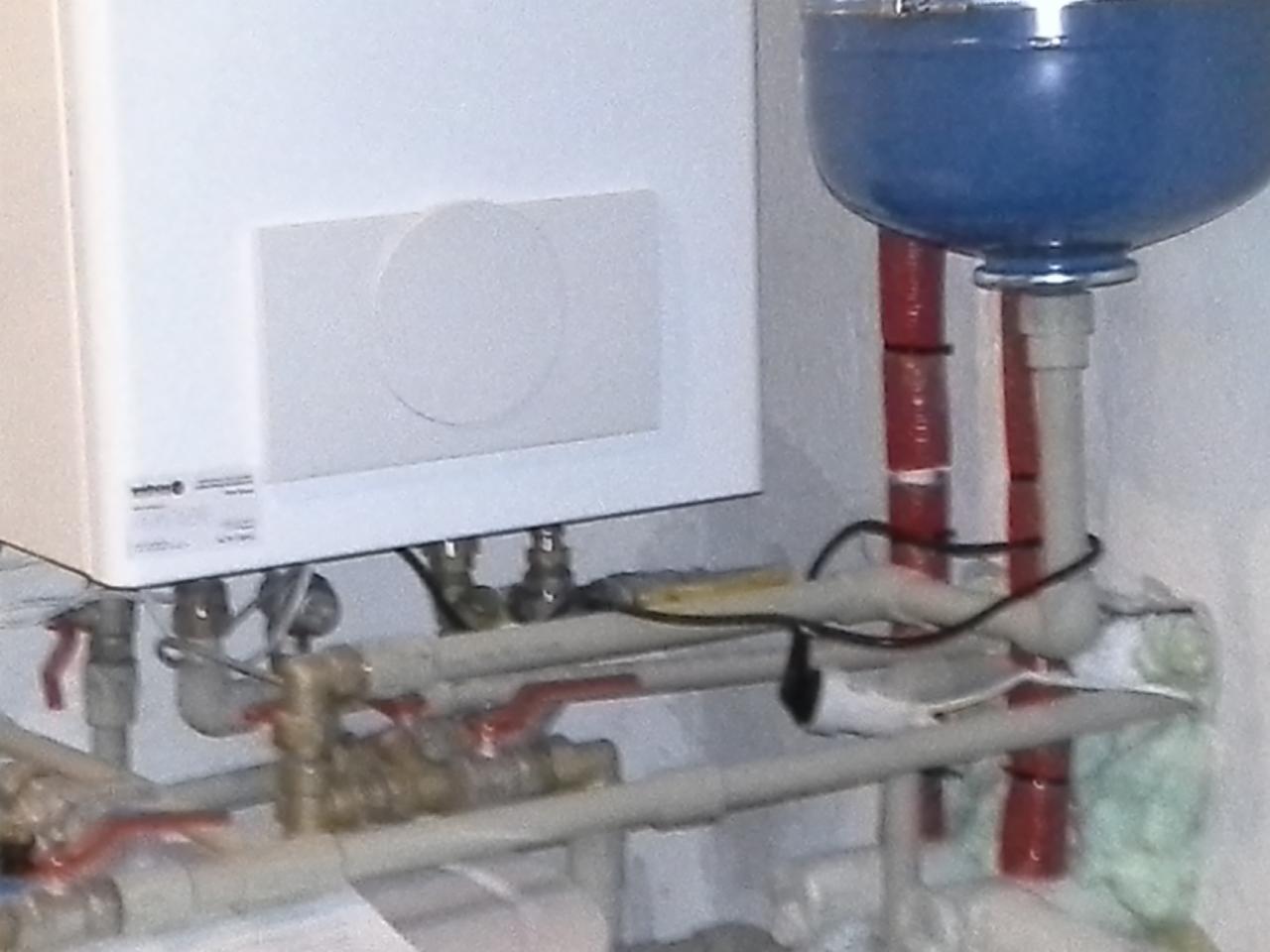Biały piec gazowy zamontowany na ścianie z widocznymi rurami, zaworami i niebieskim naczyniem przeponowym