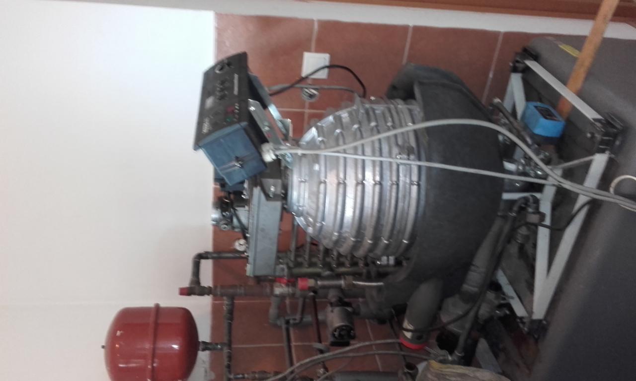 Instalacja hydrauliczna z zbiornikiem wyrównawczym, kotłem z izolacją i osprzętem, widoczne rury miedziane i sterownik, na tle ściany z brązowymi płytkami.