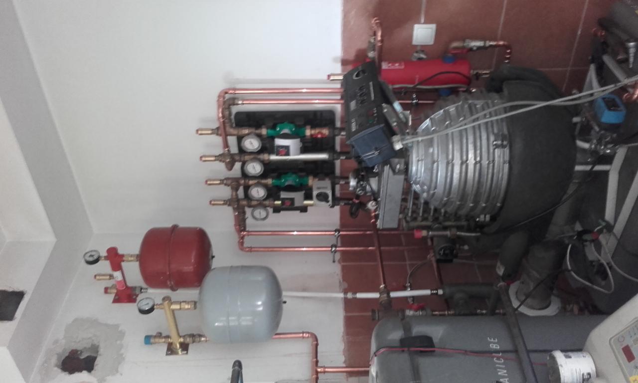 Złożona instalacja grzewcza z kotłem kondensacyjnym, zbiornikami wyrównawczymi (czerwony i szary), pompami obiegowymi i rozbudowanym systemem rur miedzianych, zamontowana w pomieszczeniu...