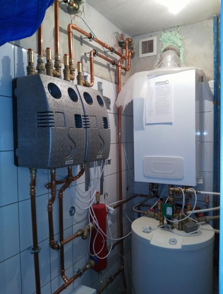 Instalacja kotła grzewczego z rozbudowaną siecią miedzianych rur i dodatkowym zasobnikiem, widoczne elementy zabezpieczone folią i pianką montażową.