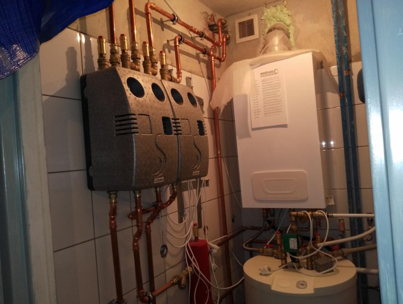 Instalacja centralnego ogrzewania z dwoma szarymi rozdzielaczami CO, miedzianymi rurami, białym piecem gazowym z dokumentacją techniczną i białym zasobnikiem ciepłej wody w małej kotłowni wyłożonej...