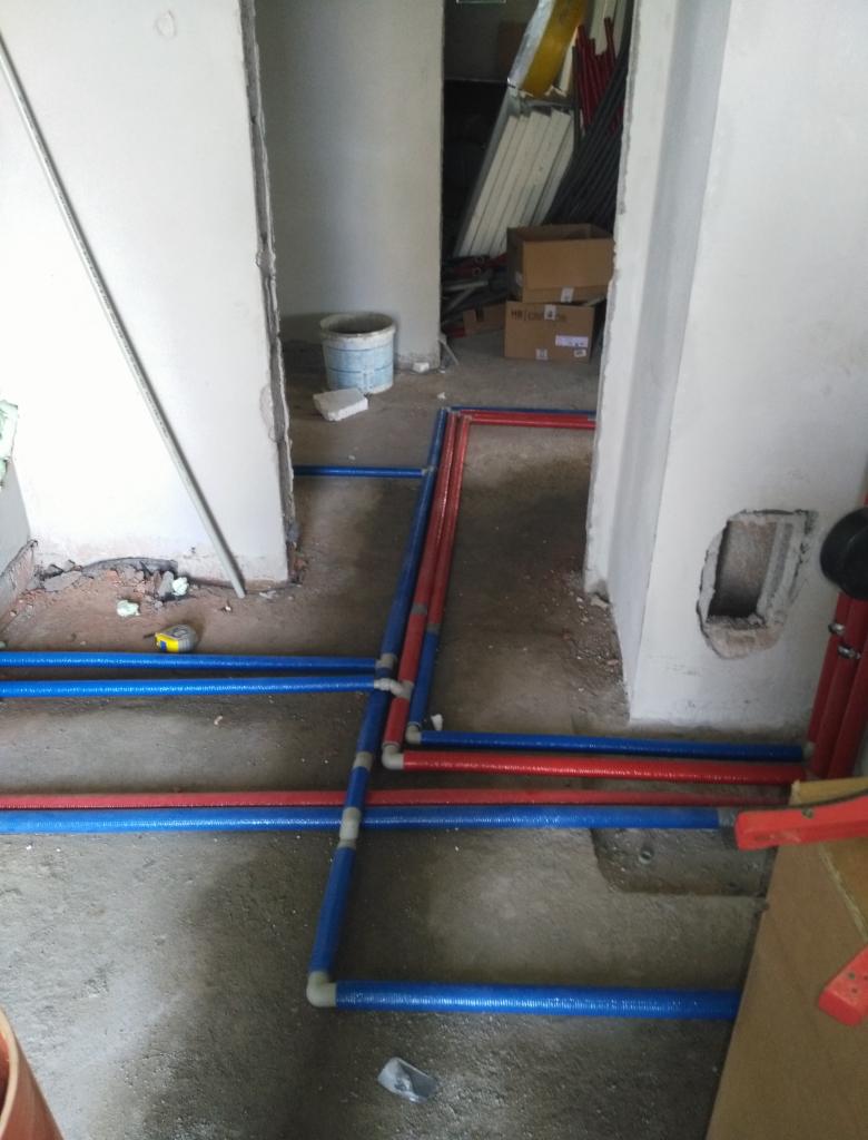 Instalacja rur grzewczych, czerwonych i niebieskich, ułożonych na surowym podłożu w trakcie remontu, widoczne połączenia i kształtki, w tle pomieszczenie z wiadrem i materiałami budowlanymi.
