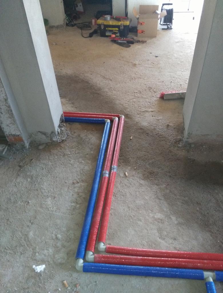 Instalacja rurowa, czerwone i niebieskie rury ułożone przy ścianie na surowym betonowym podłożu, widoczne narzędzia i kartony w tle.