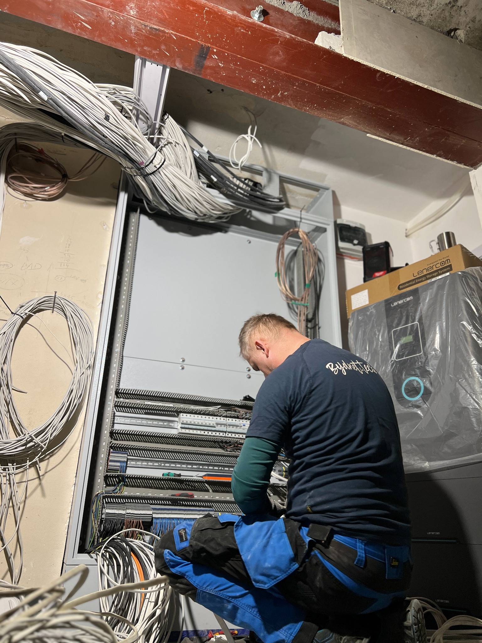 Elektryk w trakcie pracy nad rozdzielnicą elektryczną, widoczne liczne przewody, skrzynka z urządzeniem elektrycznym marki Lenercom w tle.