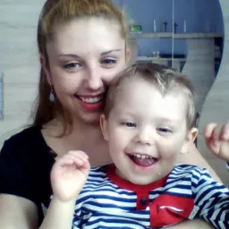 Uśmiechnięta kobieta trzymająca na rękach roześmianego chłopca w pasiastym ubraniu na tle pokoju z drewnianymi szafkami.