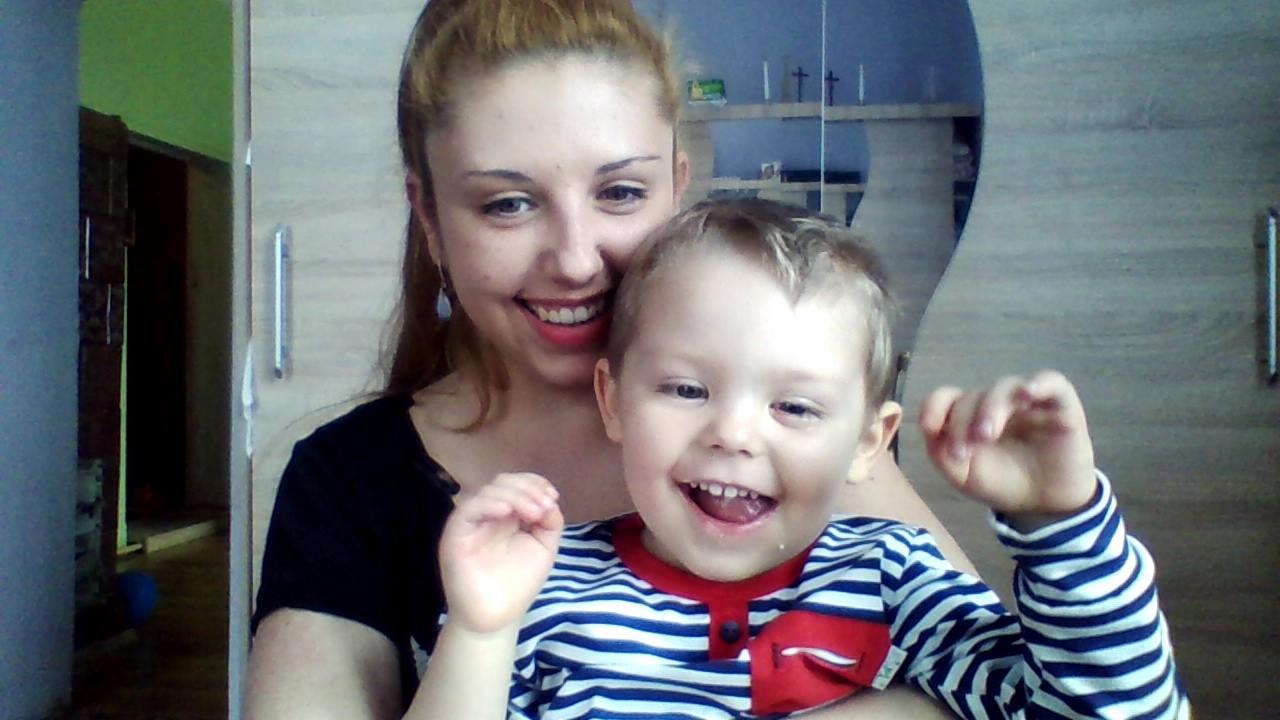 Uśmiechnięta kobieta trzymająca na rękach roześmianego chłopca w pasiastym ubraniu na tle pokoju z drewnianymi szafkami.