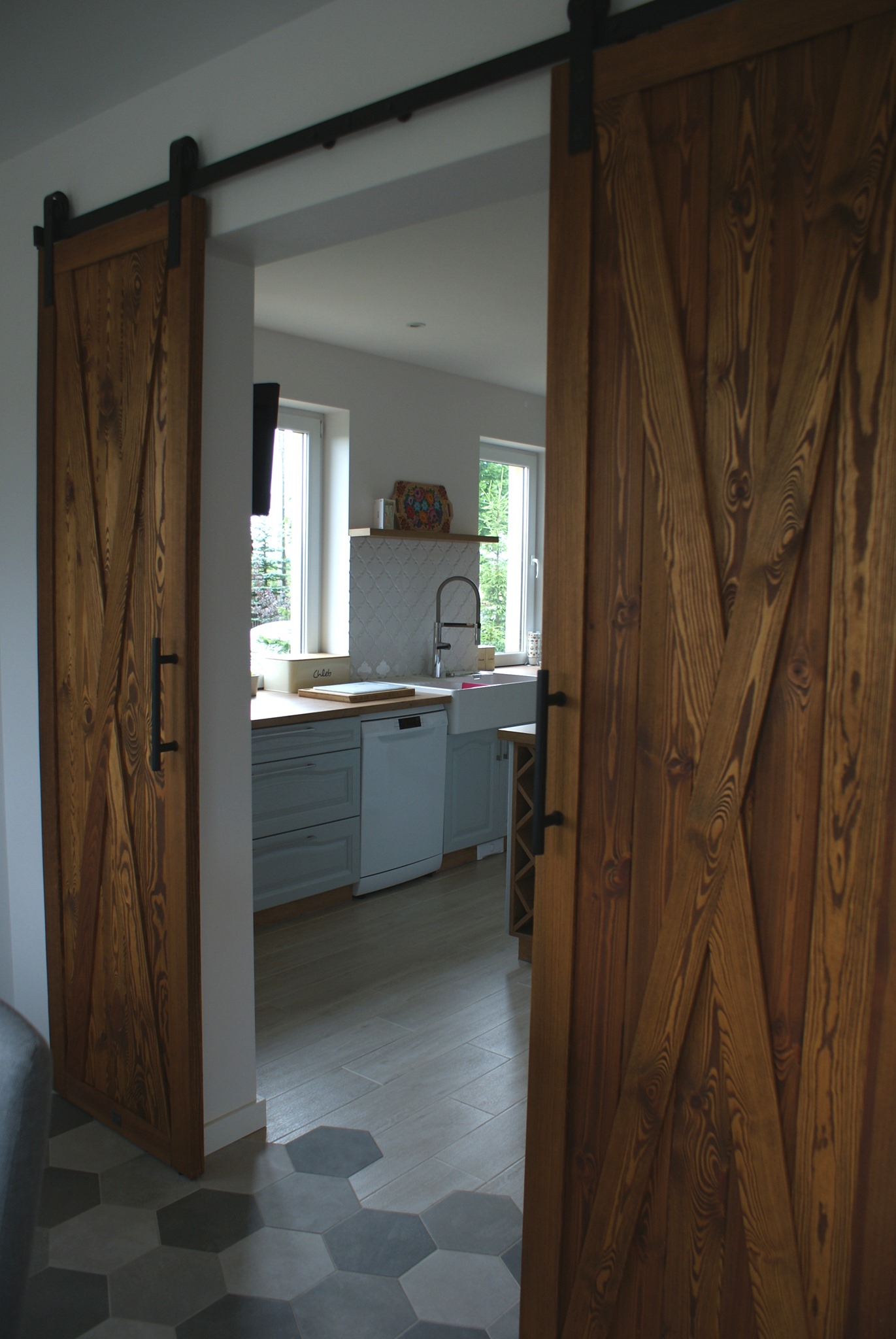 Drewniane przesuwne drzwi stodołowe z czarnymi okuciami, częściowo otwarte, prowadzące do kuchni z szarymi szafkami i białym zlewozmywakiem farmerskim, widok na podłogę z szarych płytek...