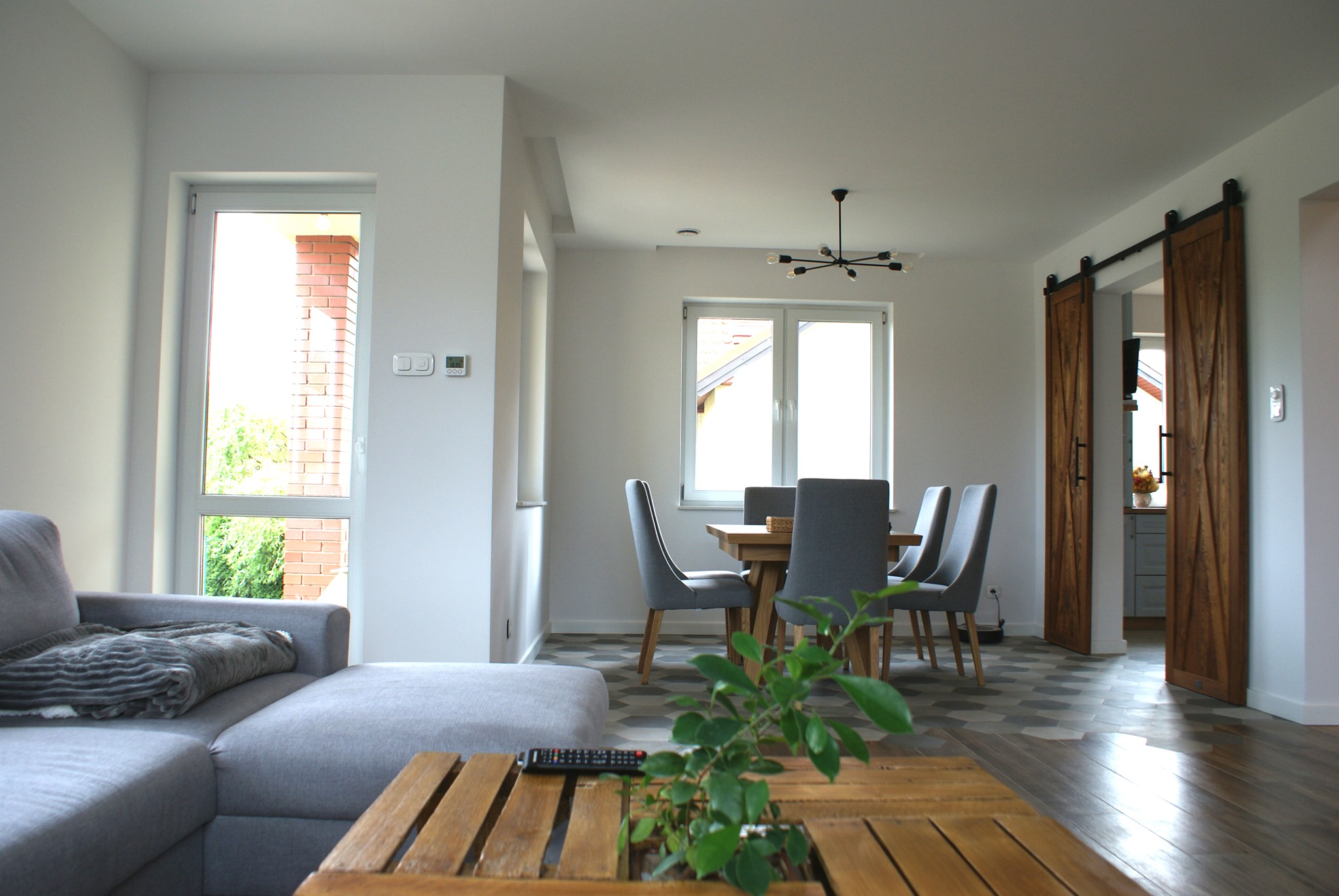 Jasne, nowocześnie urządzone wnętrze domu z szarym narożnikiem, drewnianym stolikiem kawowym z rośliną, jadalnią z szarymi krzesłami i drewnianym stołem oraz przesuwnymi drewnianymi drzwiami...