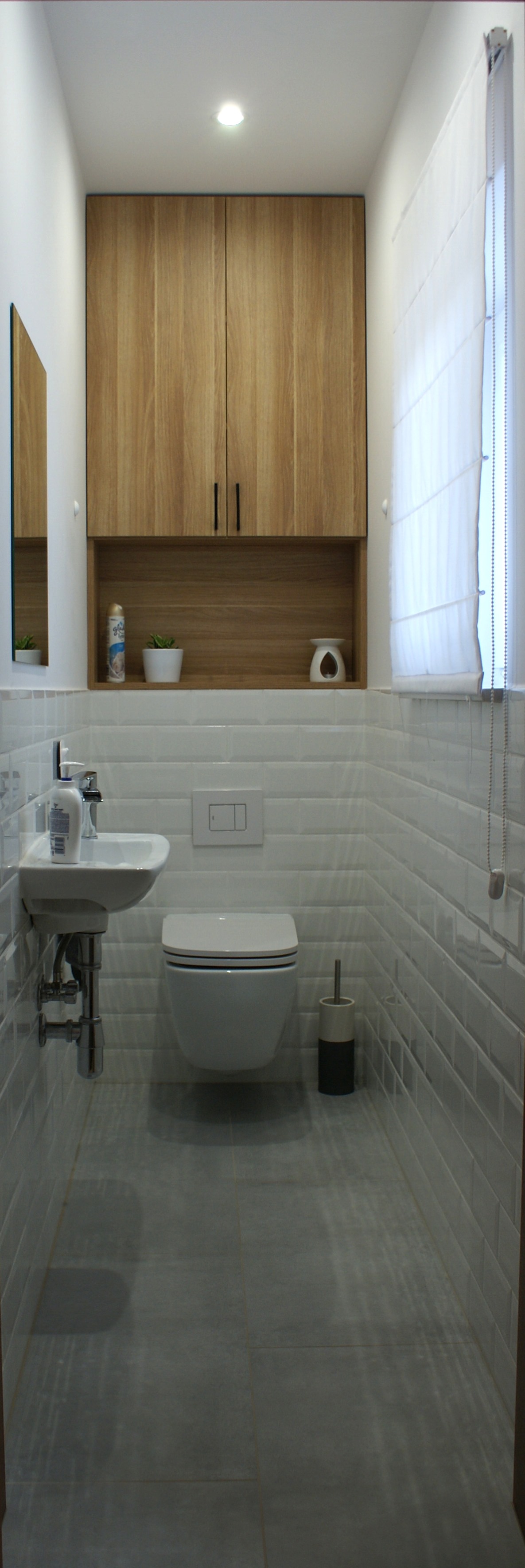 Wąska toaleta z wiszącą muszlą, umywalką narożną i szafką wiszącą z drewnianymi frontami, wykończona białymi, prostokątnymi płytkami imitującymi cegiełki.