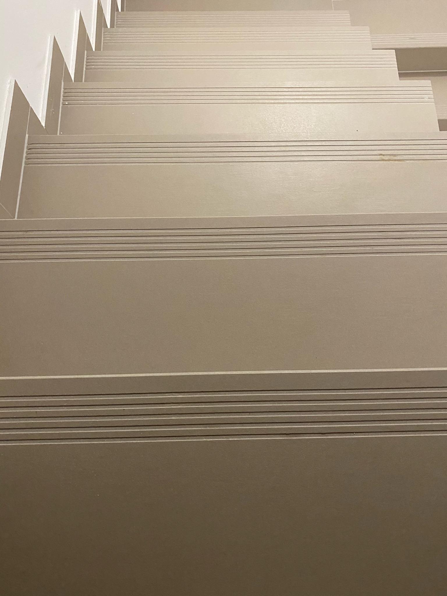 Widok z góry na beżowe schody z antypoślizgowymi rowkami, prowadzące w górę, z białym wykończeniem ściany po lewej stronie.