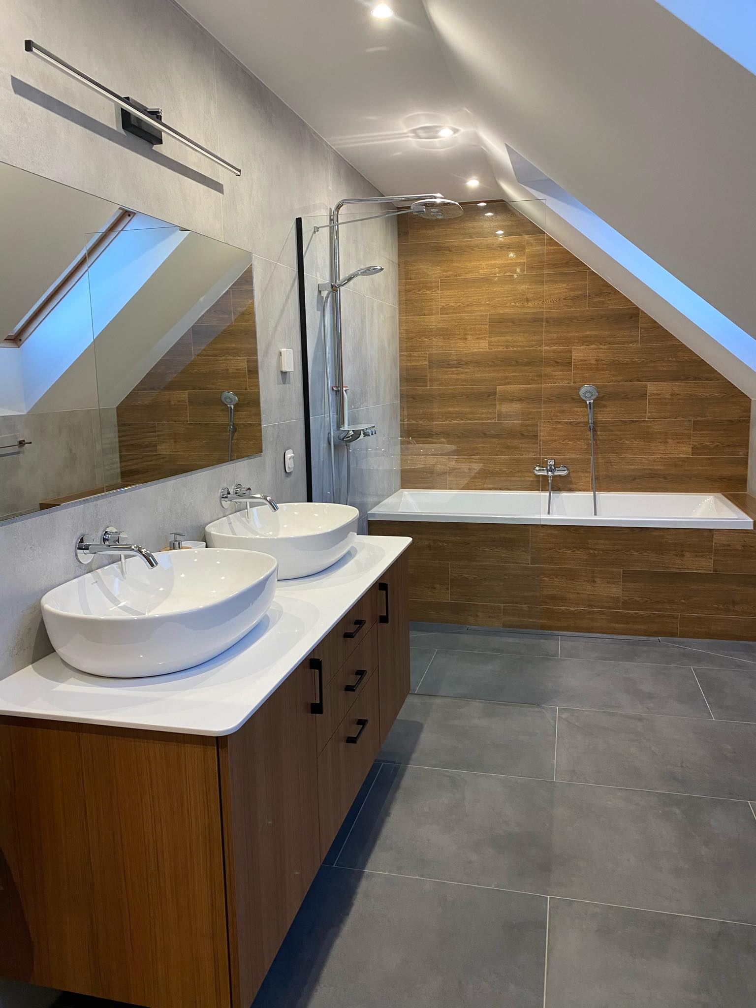 Nowoczesna łazienka na poddaszu z dwoma umywalkami nablatowymi, drewnianą zabudową szafek, brązowymi płytkami imitującymi drewno na ścianie przy wannie oraz szarą podłogą z dużych płytek.