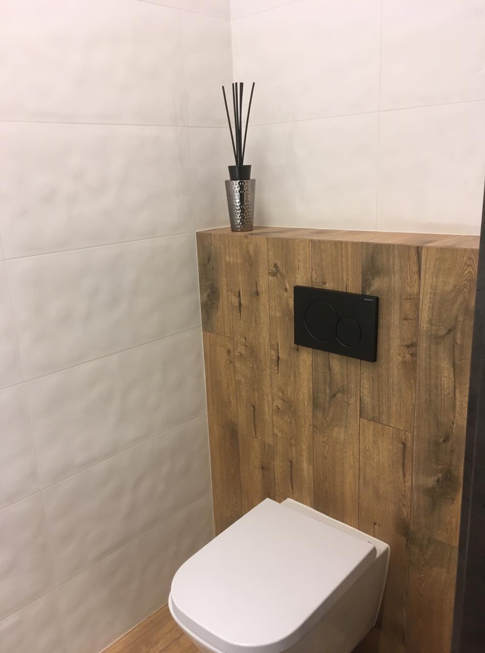 Nowoczesna toaleta z białymi płytkami i drewnopodobną obudową stelaża, czarny przycisk spłukujący, srebrny dyfuzor zapachowy z czarnymi patyczkami.
