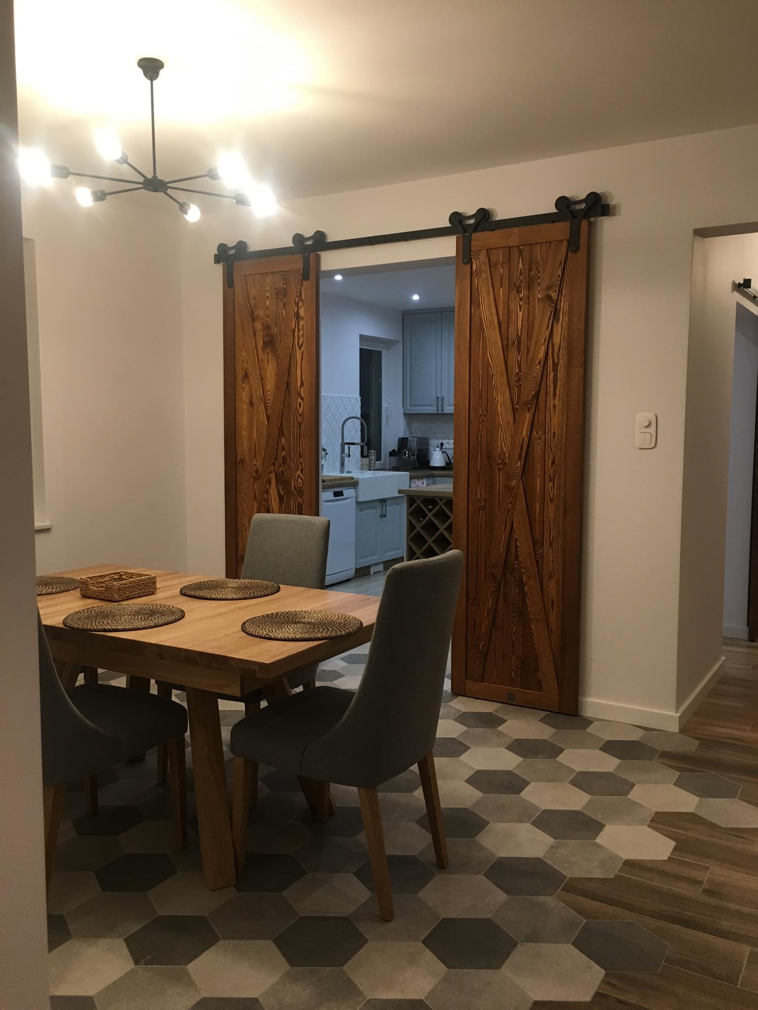 Drewniane przesuwne drzwi w stylu rustykalnym, z czarnymi okuciami, oddzielające jadalnię od kuchni z szarymi szafkami i białym zlewem farmerskim; na stole ustawione podkładki i koszyk.