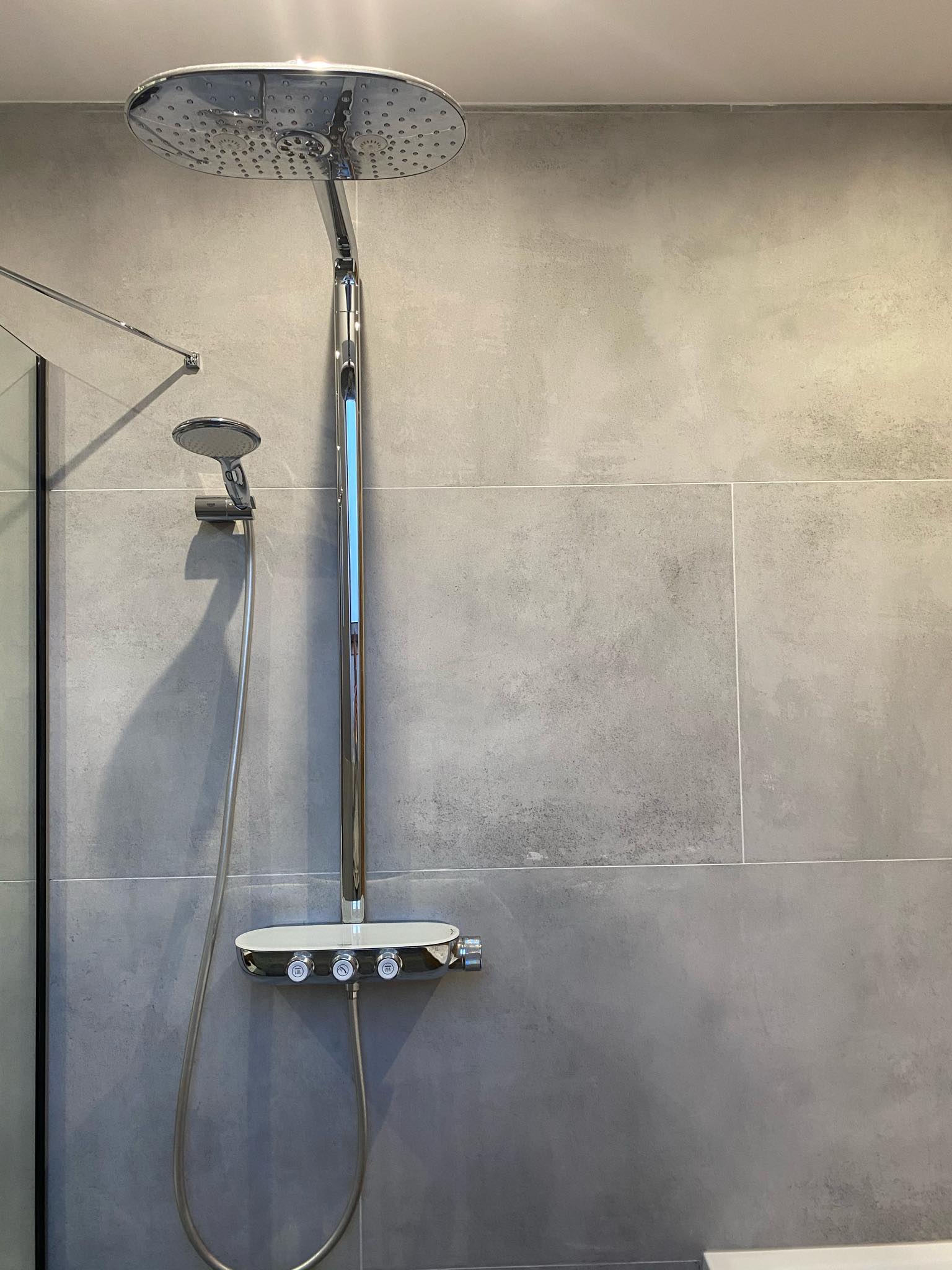 Nowoczesny zestaw prysznicowy z deszczownicą i słuchawką, zamontowany na ścianie wyłożonej szarymi płytkami o fakturze betonu.