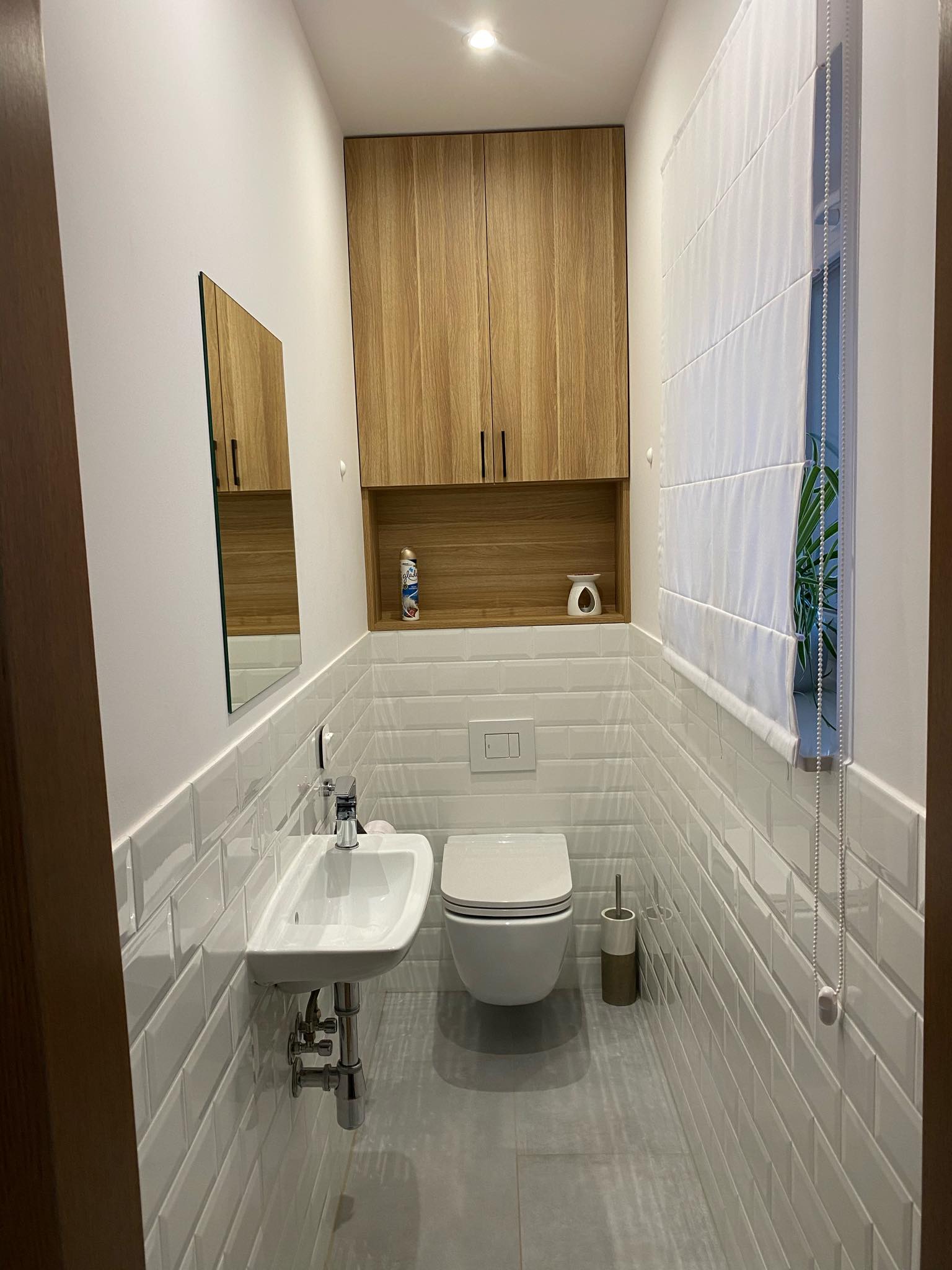 Wąska toaleta w jasnych barwach z białymi, prostokątnymi płytkami na ścianach, podwieszana szafka z jasnego drewna nad zabudowaną spłuczką, mała umywalka z chromowaną armaturą i szarą podłogą.