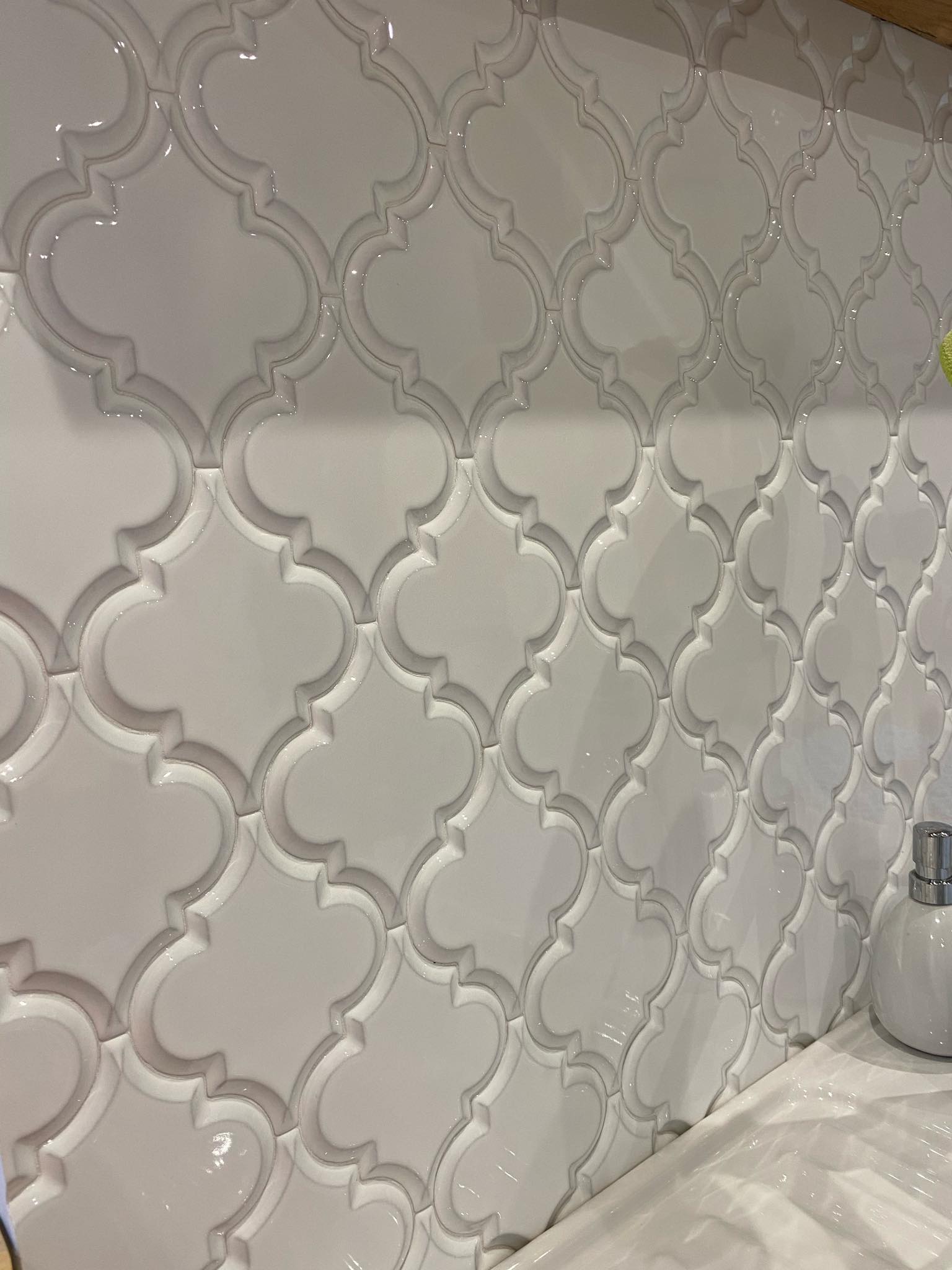 Białe, błyszczące płytki w marokański wzór ułożone na ścianie nad umywalką, widoczny dozownik do mydła.