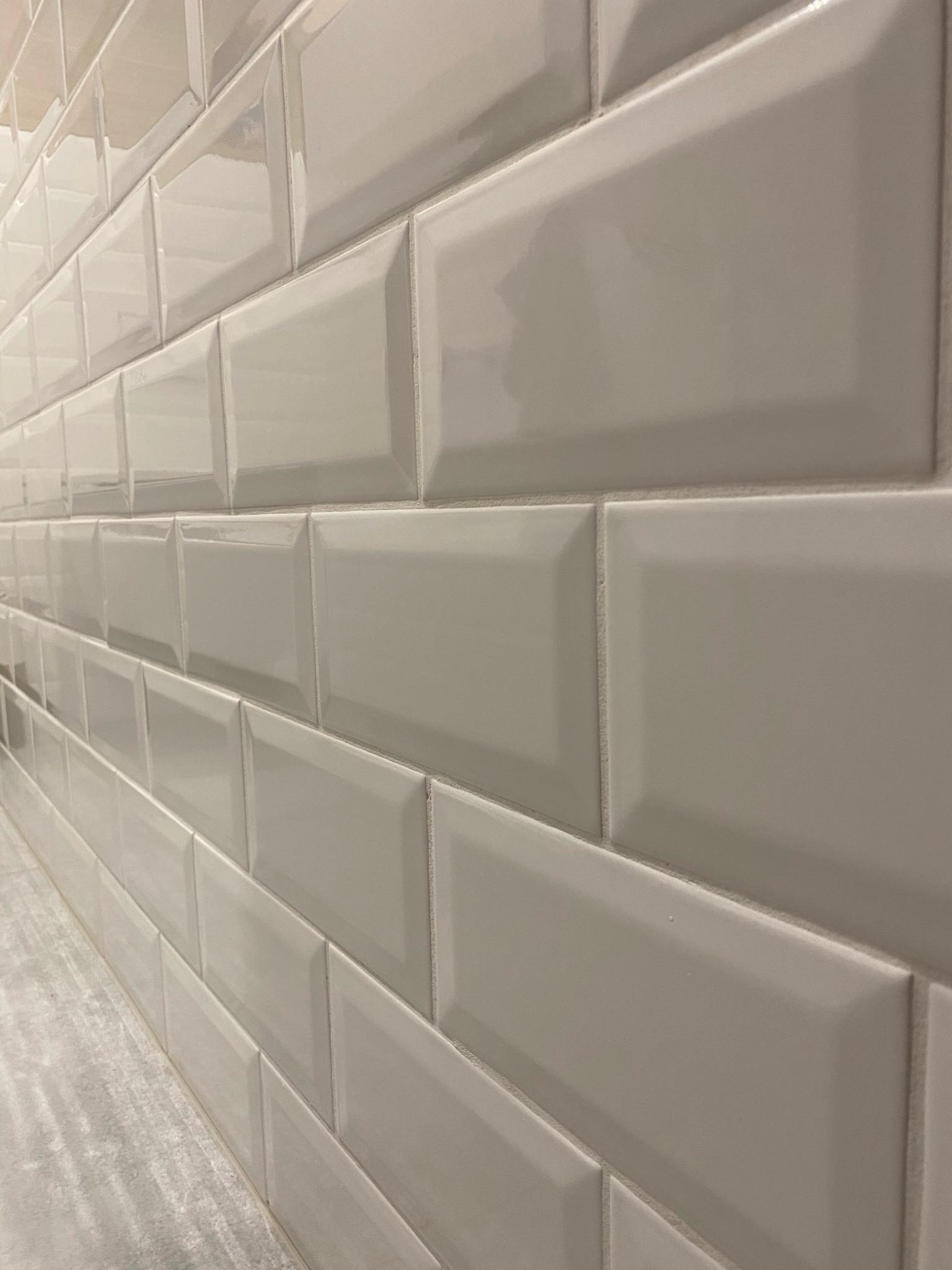 Ściana wyłożona błyszczącymi, beżowymi płytkami typu metro z widocznymi fugami. Płytki ułożone poziomo, z delikatnym efektem 3D. Widoczny fragment blatu pod płytkami.
